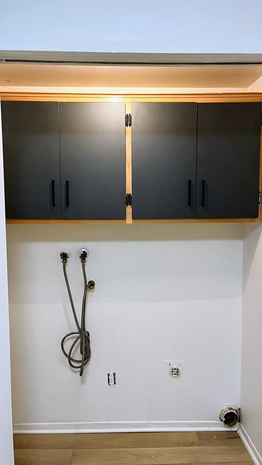 Empty laundry area with dark gray upper cabinets, plumbing connections, electrical outlet, and vent pipe on a white wall.
