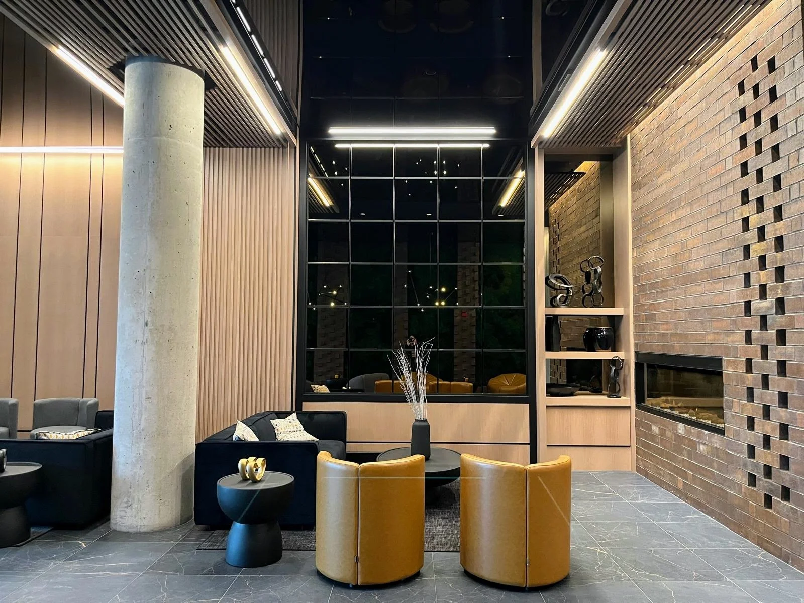 Modern hotel lobby with black and mustard chairs, a round black coffee table, decorative sculptures on a shelf, and a brick wall with a horizontal fireplace.
