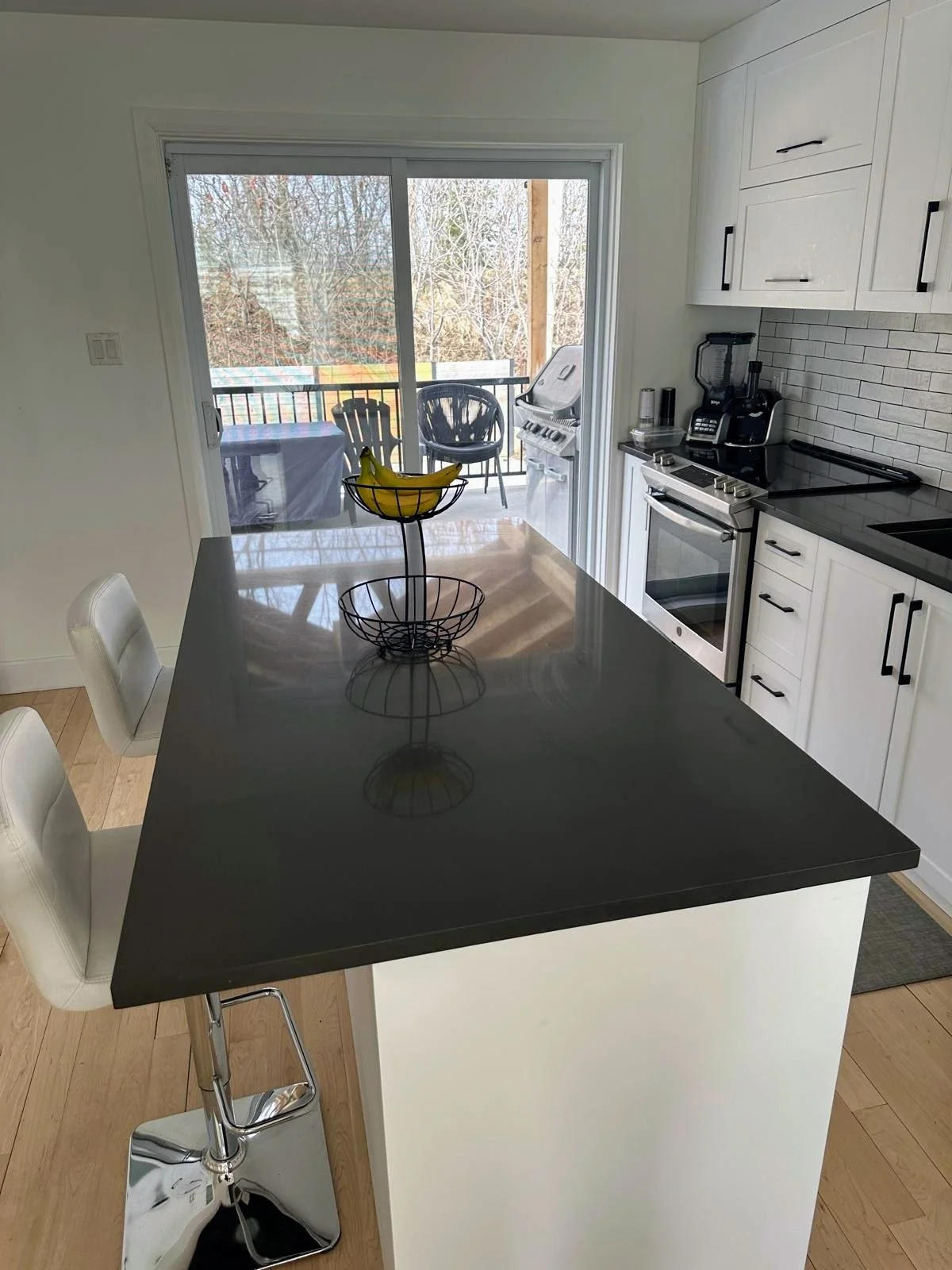 Modern kitchen with white cabinets, black countertops, and a black island. There are two white chairs at the island. A sliding glass door leads to an outdoor patio with a table, chairs, a grill, and trees outside.
