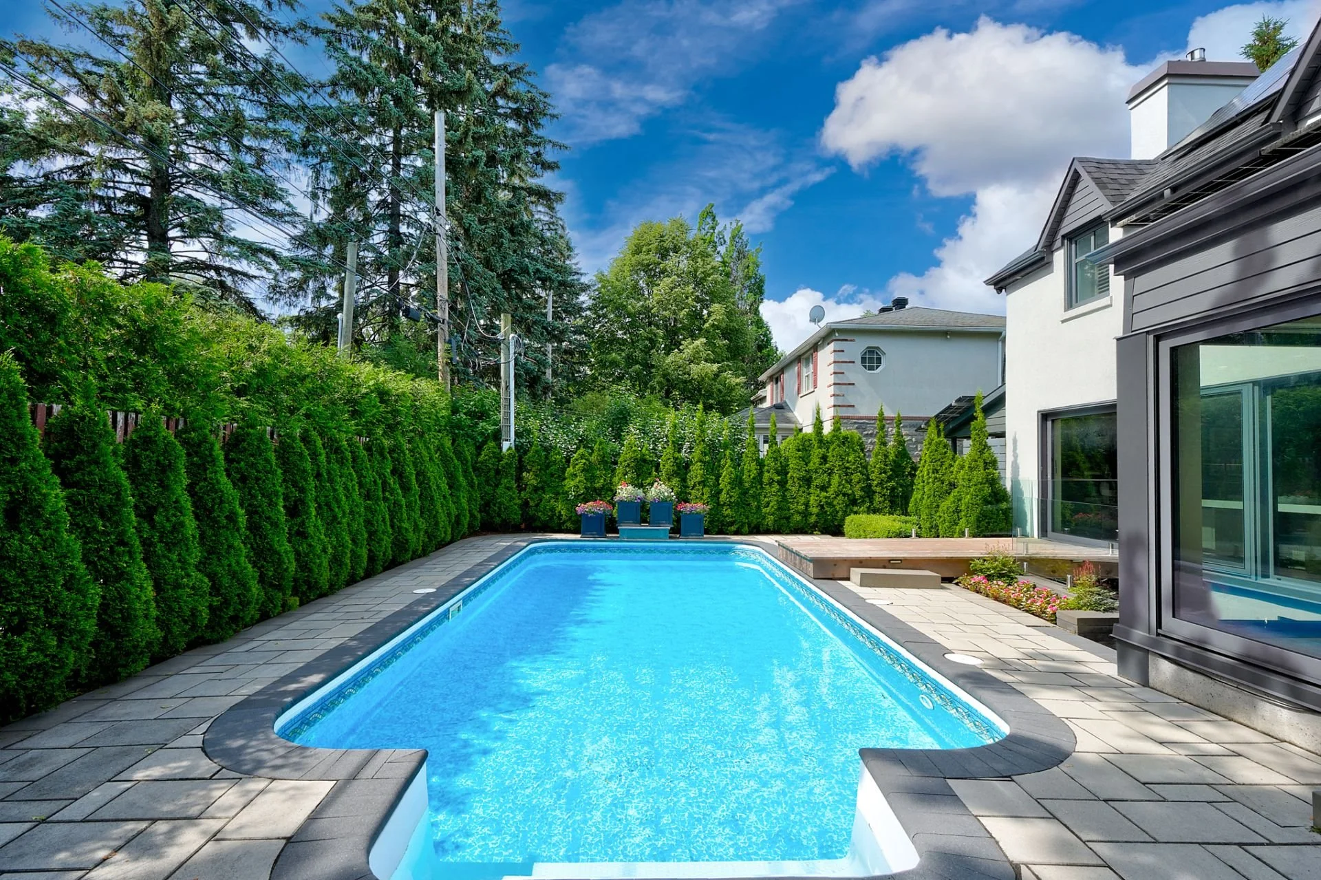 A backyard with a rectangular in-ground swimming pool, surrounded by gray stone pavers, enclosed by tall green hedges and trees, with a house featuring large windows and neighboring houses in the background, under a partly cloudy blue sky.