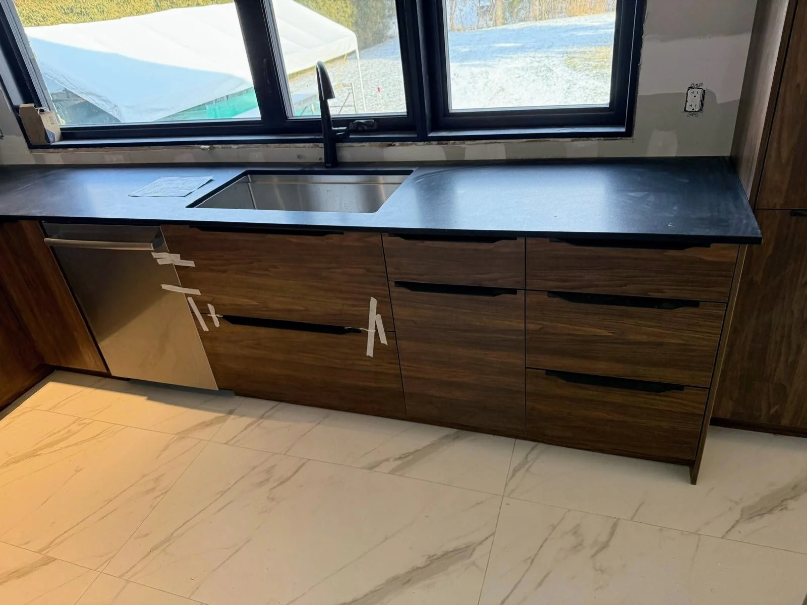 Kitchen cabinet with dark wood finish, black countertop, stainless steel sink, black faucet, large window above the sink, and an electrical outlet on the wall. The cabinet has several drawers and doors, some with handles.