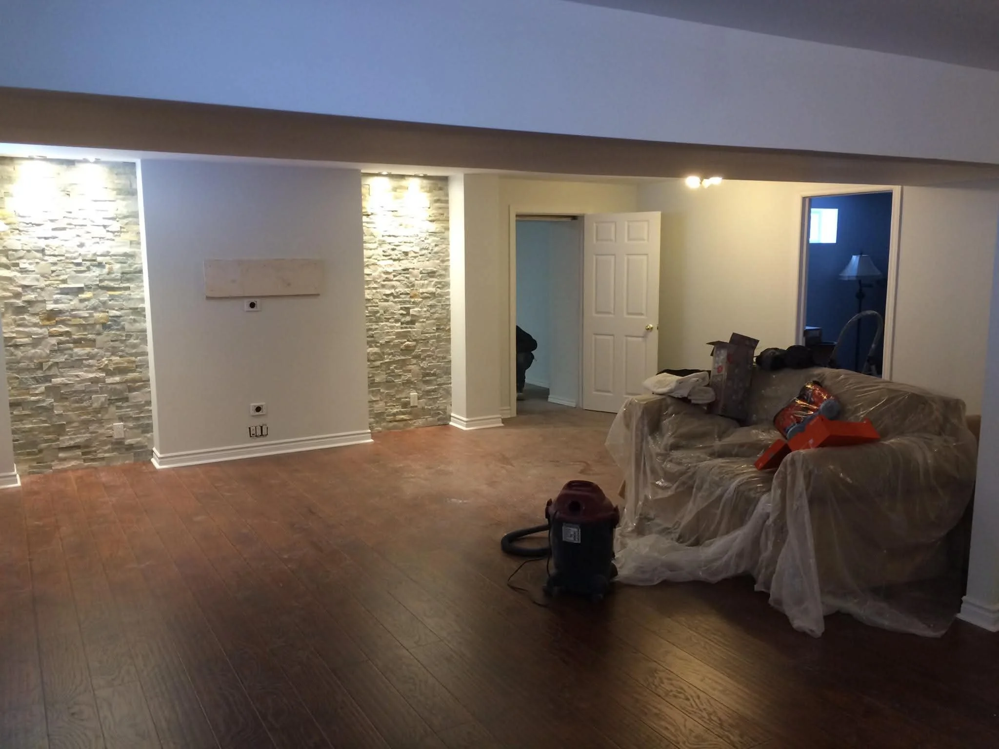 Living room under renovation with hardwood flooring, a stone accent wall, and a plastic-covered sofa with tools and supplies on it.
