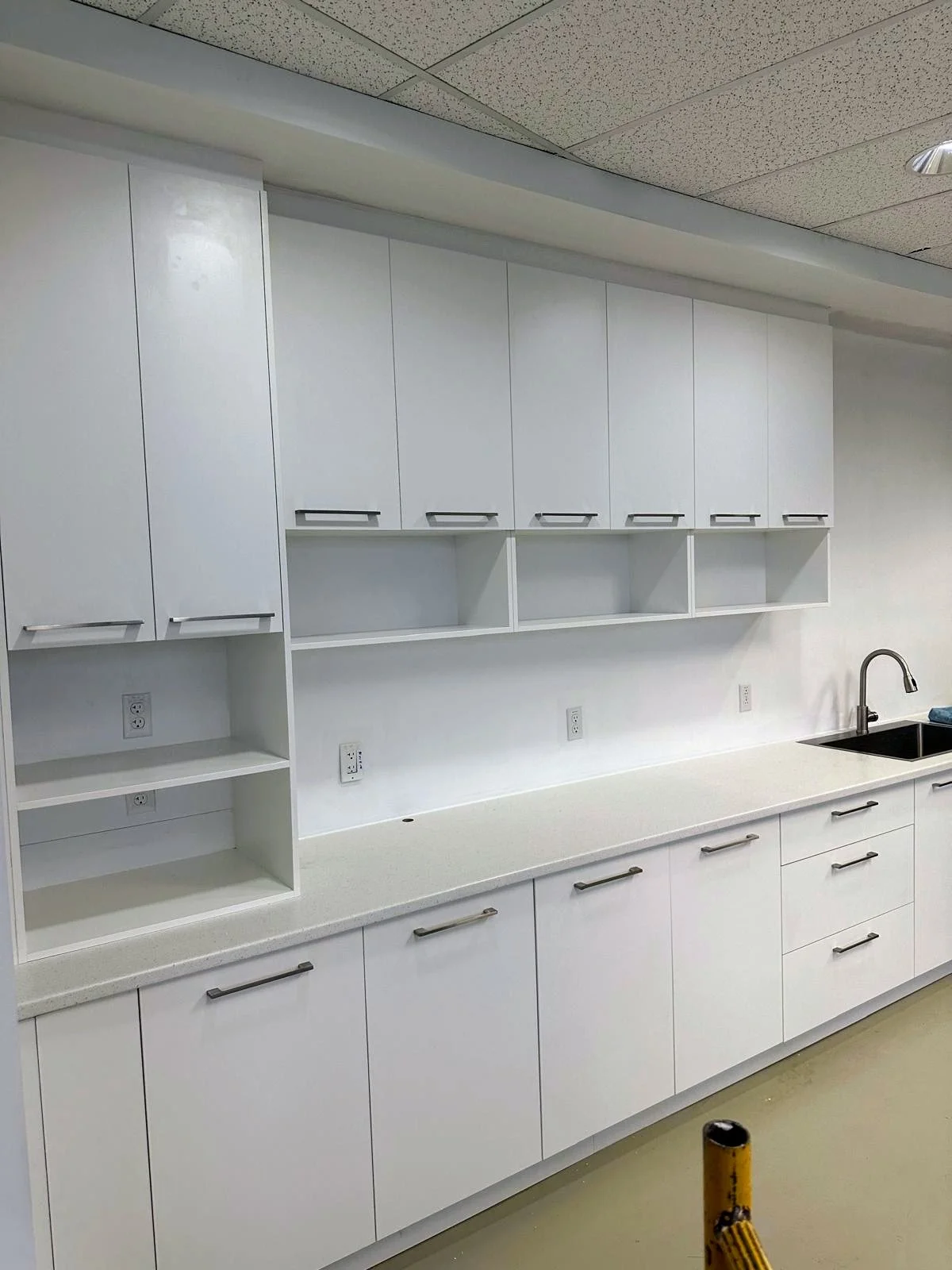 Empty white kitchen cabinets and countertop with a stainless steel sink and faucet, multiple electrical outlets, and a drop ceiling, in a clean and minimalist style.