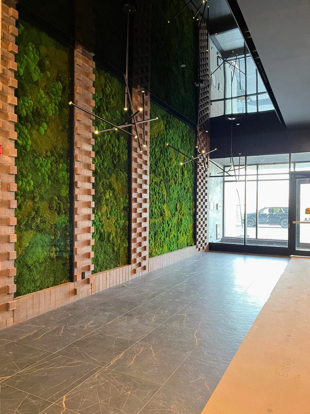 Interior of an upscale building with a wall decorated with green moss and brick patterns, modern black chandeliers, and a large glass window at the front entrance showing a parked car outside.