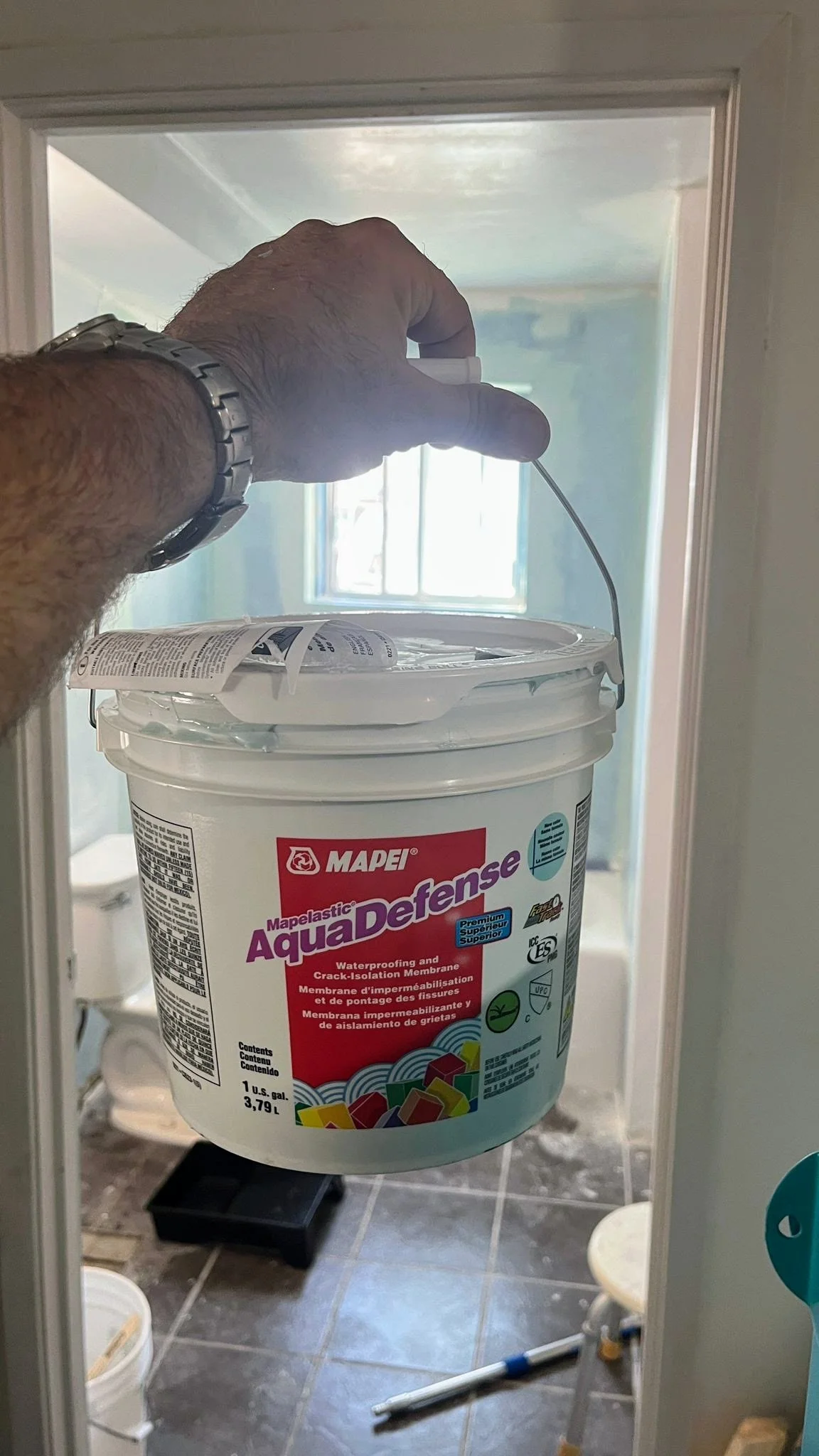 A person holding a paintbrush with a bucket of Mapelastic AquaDefense waterproofing membrane, partially visible in a bathroom with a toilet, stool, and tools on the floor.
