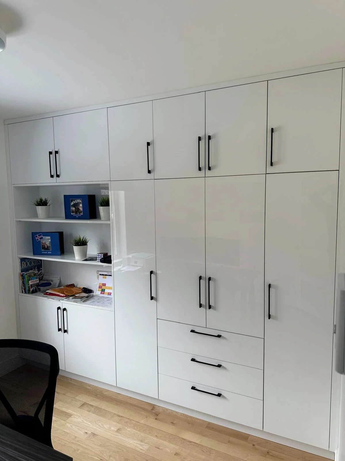 White built-in storage cabinets with black handles, a bookshelf with books and decorative items, and a wooden floor.