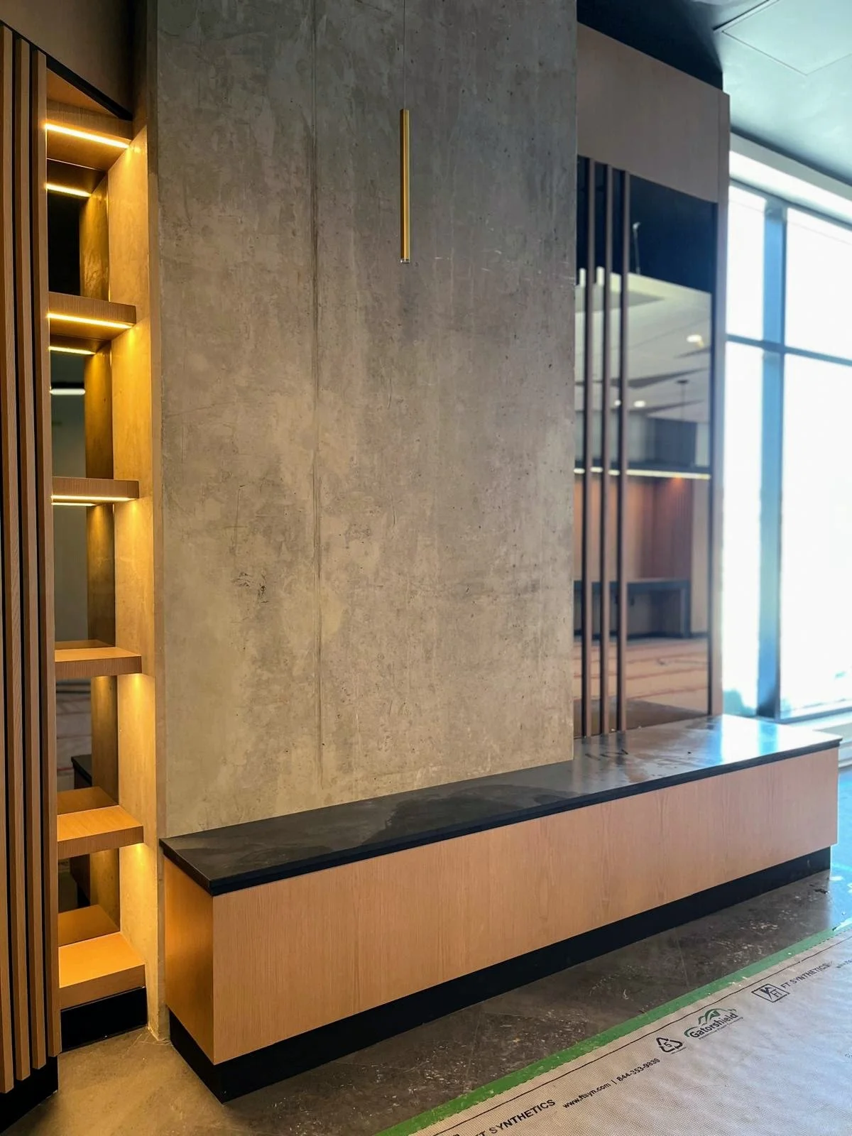 Modern interior space with a beige concrete wall, wooden shelving with lighting, and a wooden bench with a black marble top near large windows.