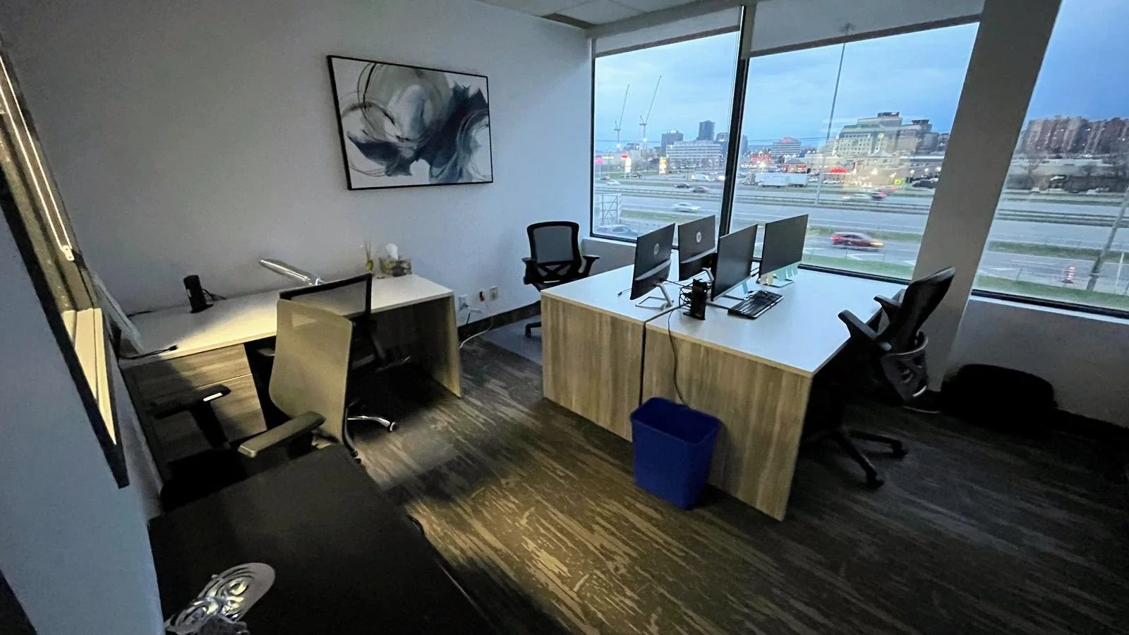 Office with four computer monitors on a desk near large windows overlooking a city highway at dusk.