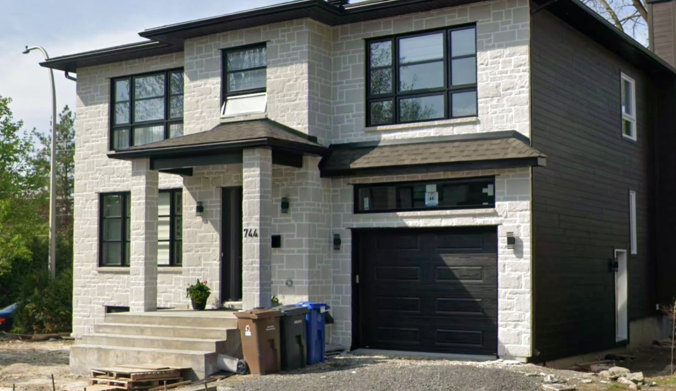 Modern two-story house with white brick facade, dark window frames, and a black garage door. Front porch with steps and a potted plant. Construction materials and trash bins in front of the house.