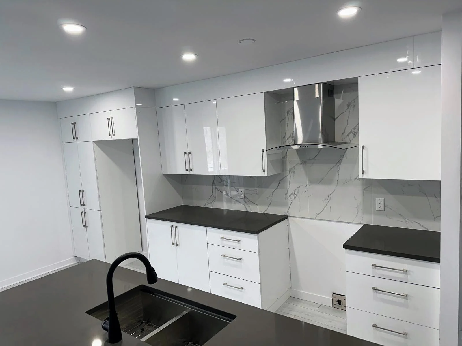 Modern kitchen with white cabinets, black countertops, marble backsplash, stainless steel range hood, and black faucet over sink.