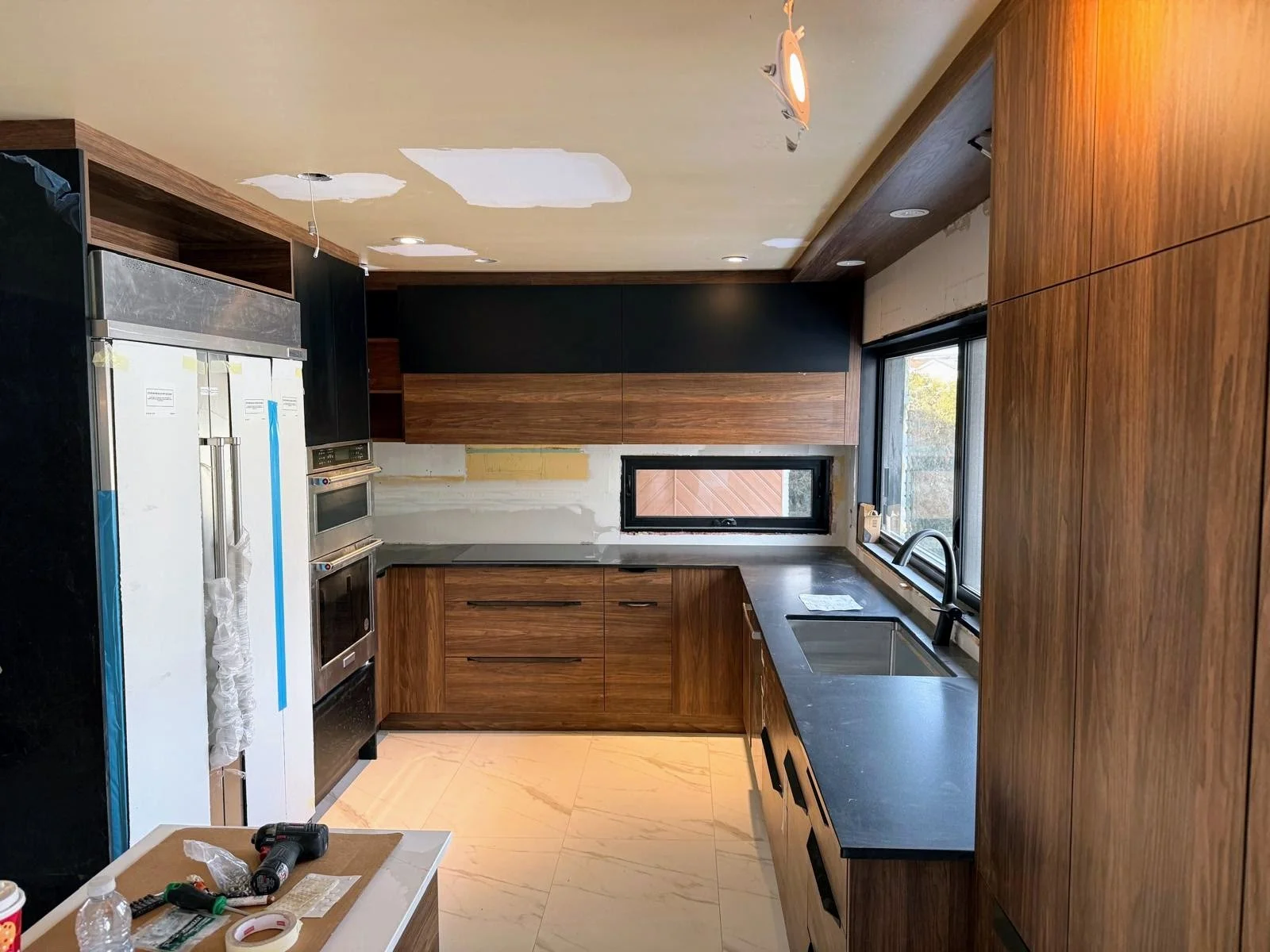 Modern kitchen under construction with wooden cabinets, black countertops, stainless steel appliances, a window above the sink, and construction tools on the countertops.
