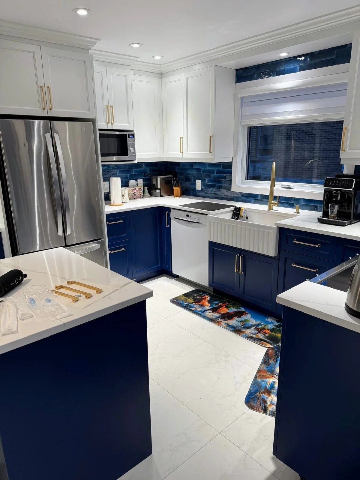 Modern kitchen with white upper cabinets, blue lower cabinets, blue backsplash, stainless steel refrigerator, microwave, dishwasher, and black coffee maker. Window above a farmhouse sink, with some mats on the floor.