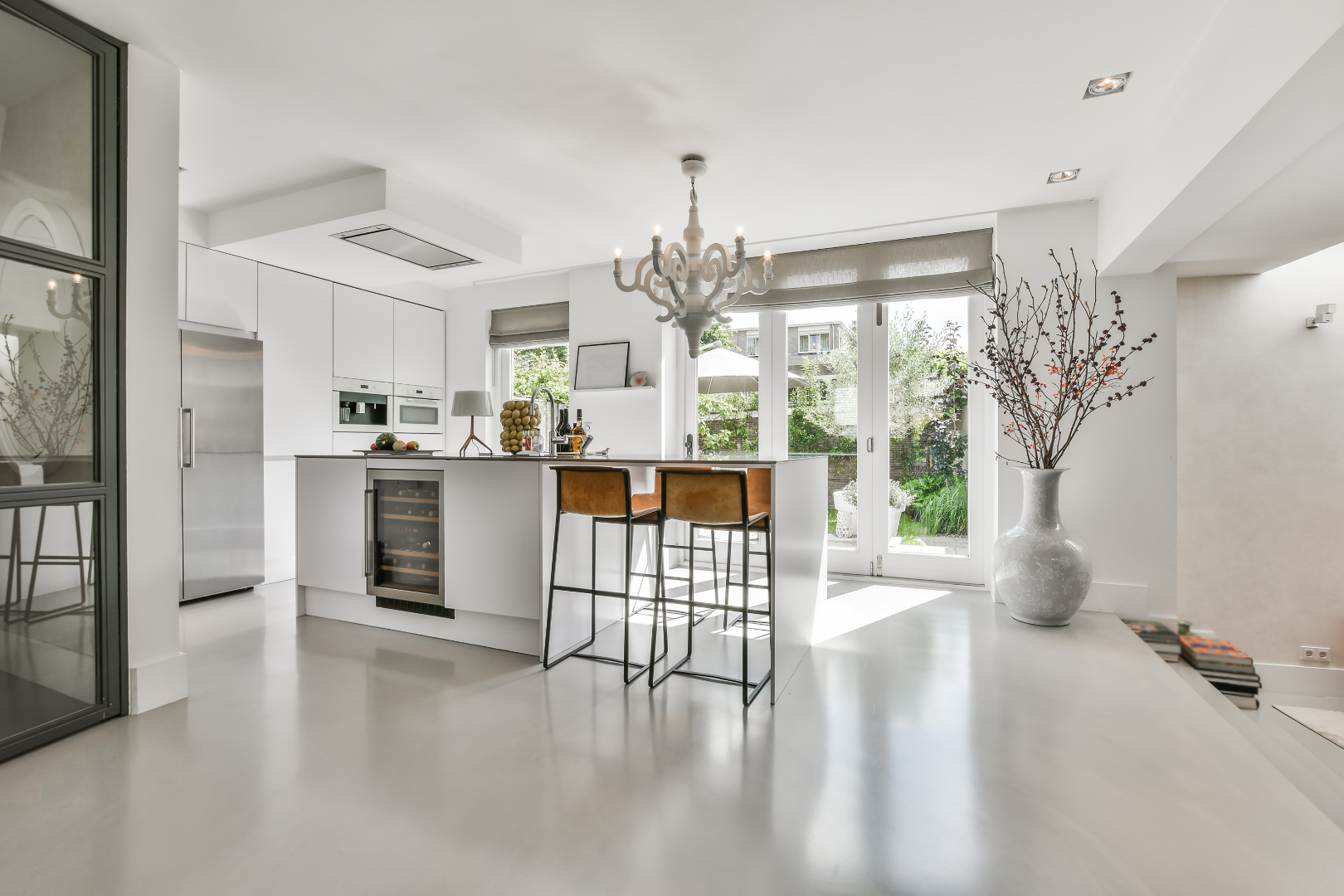 Modern white kitchen with island, brown barstools, large vase with branches, and sliding glass doors to a garden.