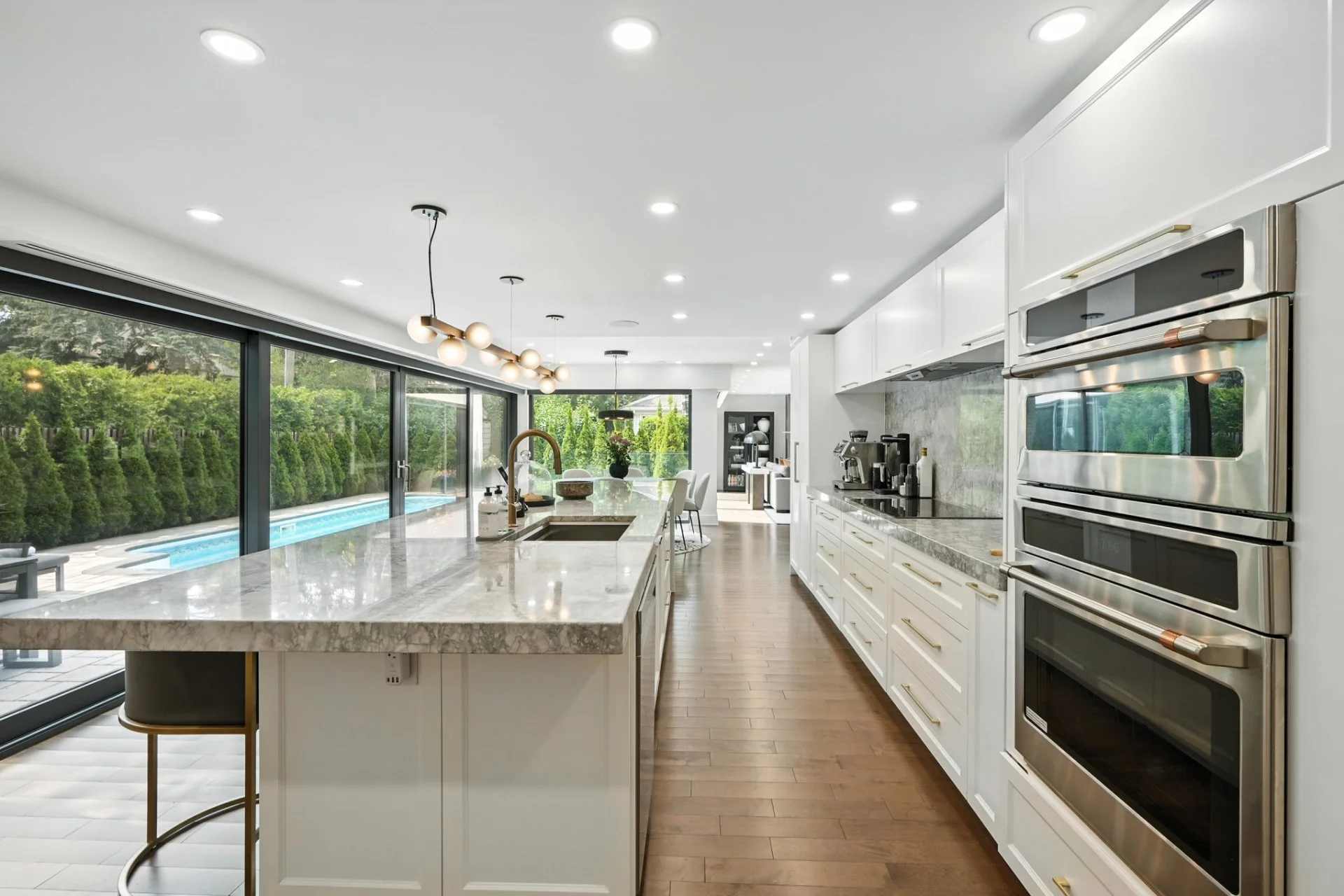 Modern white kitchen with marble countertops, stainless steel appliances, and large glass sliding doors leading to an outdoor pool area.