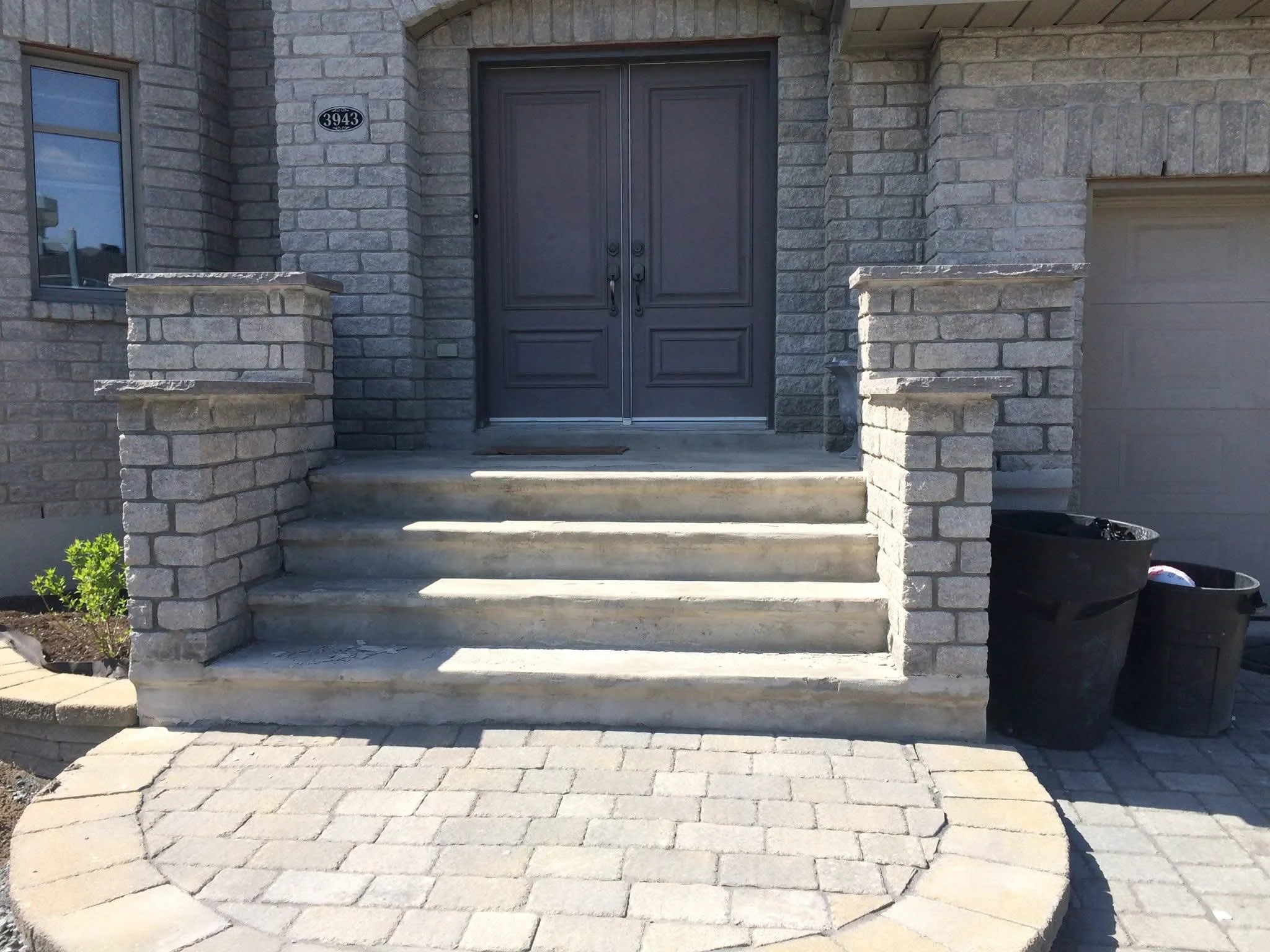 Front entrance of a house with brick steps, brick pillars, a dark double door, window on the left, and garbage bins to the right.