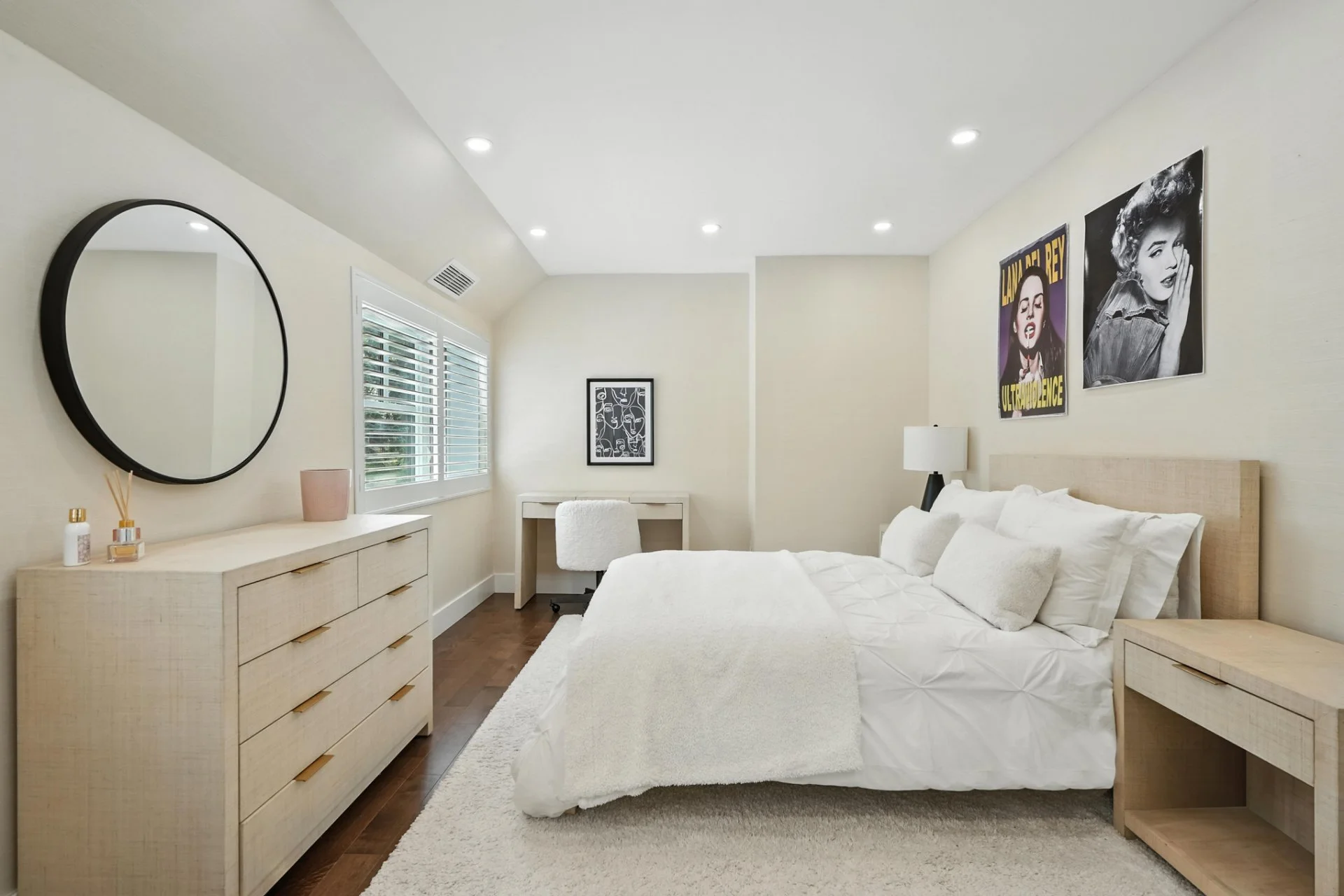 A modern bedroom with beige walls, a large window with white shutters, a bed with white bedding and pillows, two nightstands, a dresser with a round mirror, a small desk with a chair in the corner, and wall art including black and white photos and co