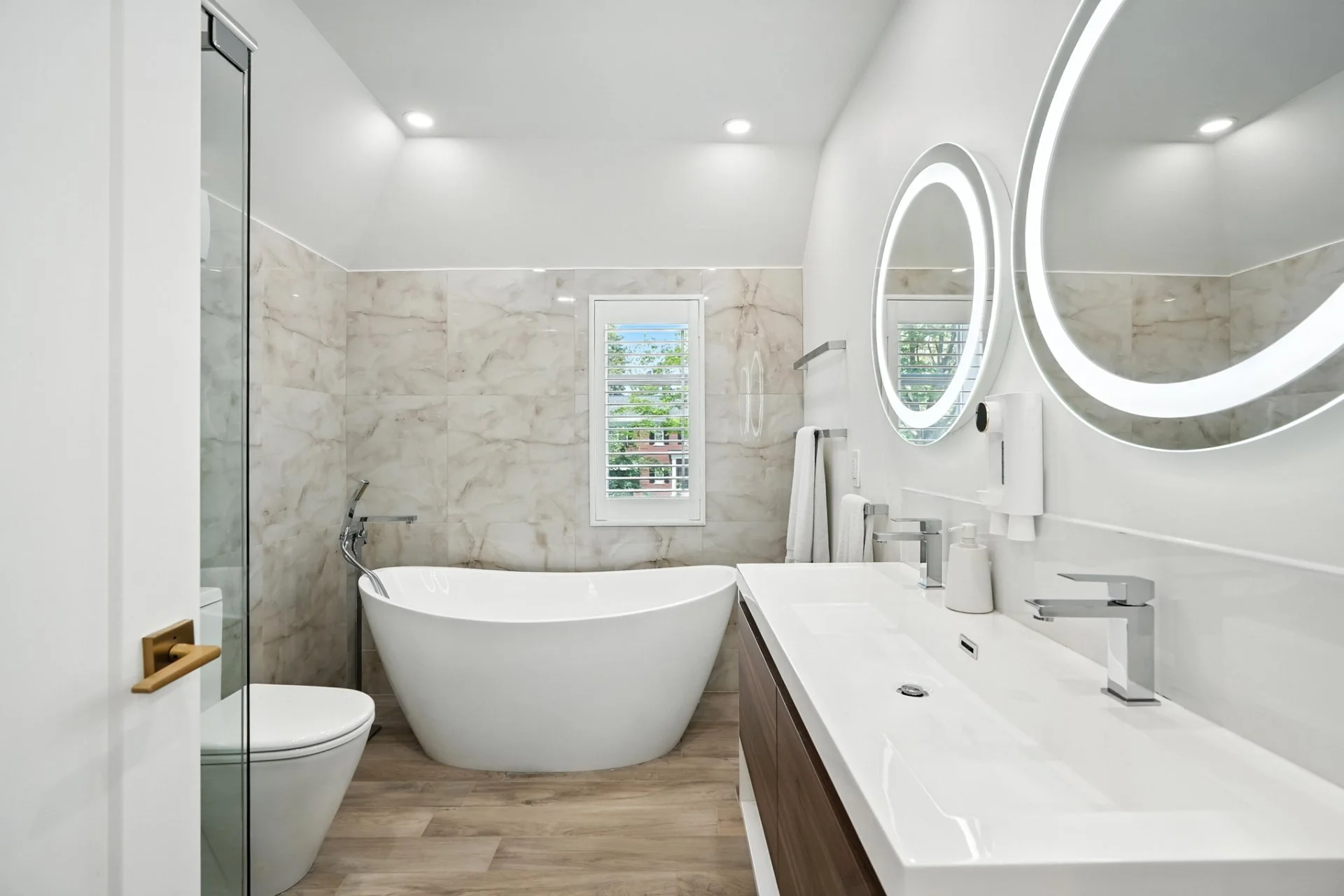 Modern bathroom with white bathtub, toilet, dual sinks, round illuminated mirrors, towel racks, wood vanity, marble wall tiles, and small window showing greenery outside.