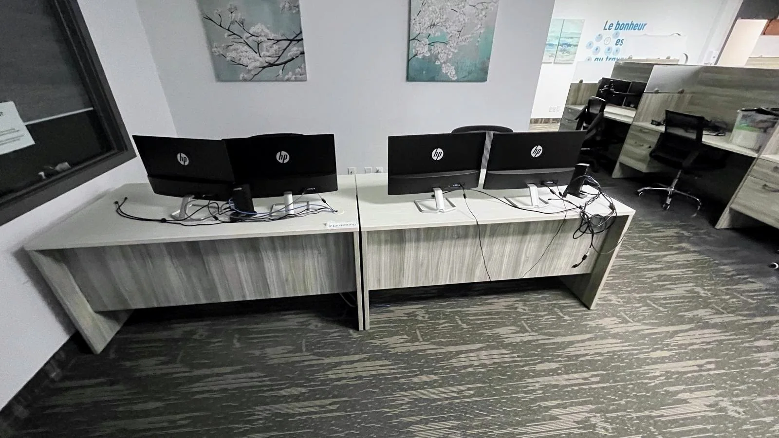 Office workspace with three HP computer monitors on a shared desk, chairs, wall art of cherry blossoms, and additional workstations in the background.
