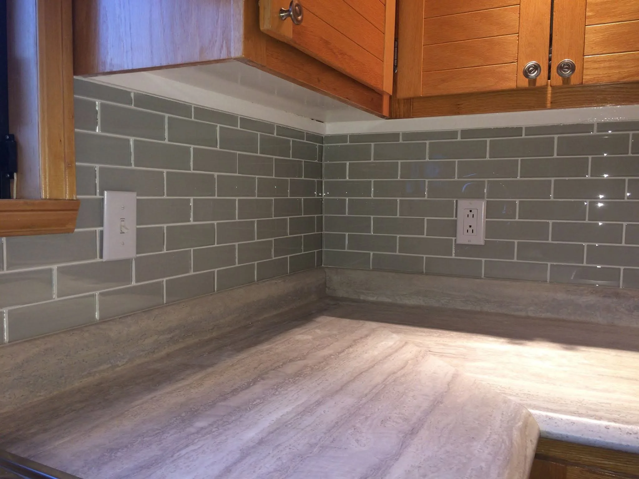 Kitchen countertop with gray granite, dark green subway tile backsplash, and wooden cabinets above. Two electrical outlets on the backsplash.