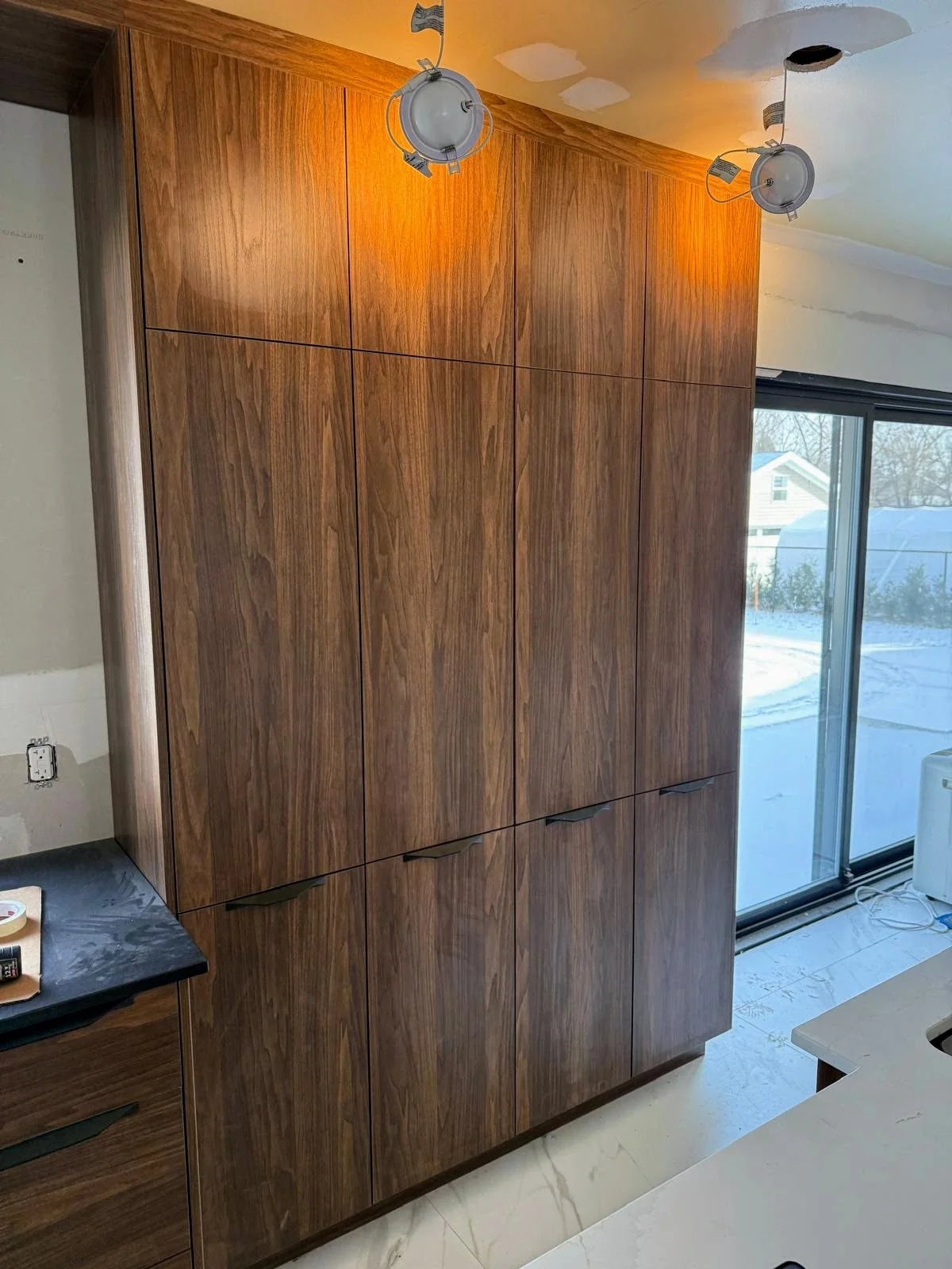 Large wooden cabinet or closet in a room under construction, with visible hardware and track lighting on the ceiling, next to a sliding glass door leading to a snowy outdoor area.