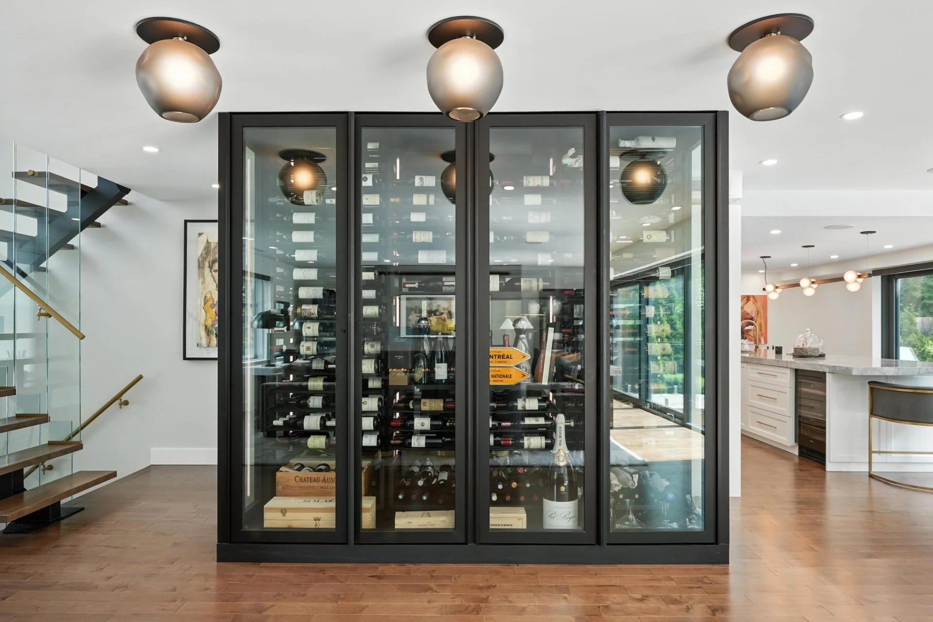 Modern wine cellar with glass door, filled with wine bottles on racks, located in a contemporary home with wood flooring, adjacent to a staircase and a kitchen area.