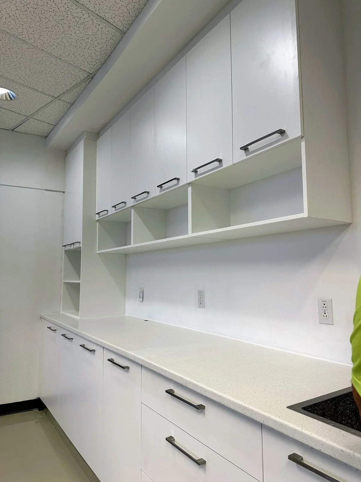 White kitchen cabinets with black handles above and below a light-colored countertop, with electrical outlets on the wall and a stainless steel sink in the corner.