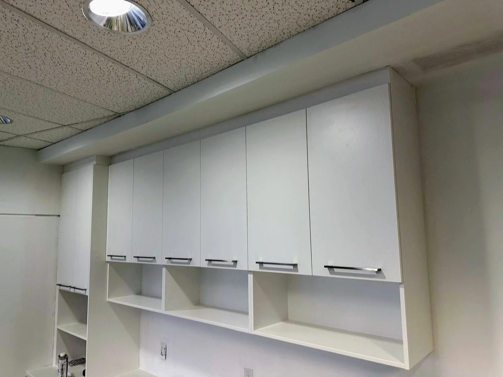 A wall of white cabinets with black handles above a white counter in a room with a textured drop ceiling and recessed lights.