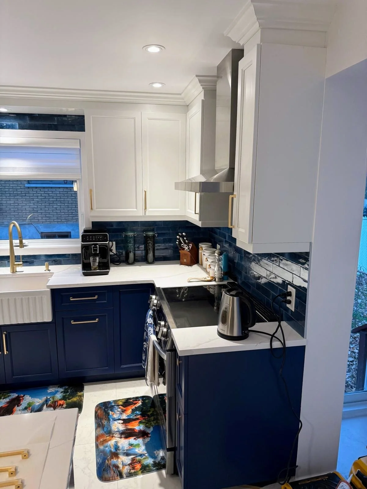 A modern kitchen with white upper cabinets, blue lower cabinets, a black and white patterned rug, and a window with a view of a brick building.