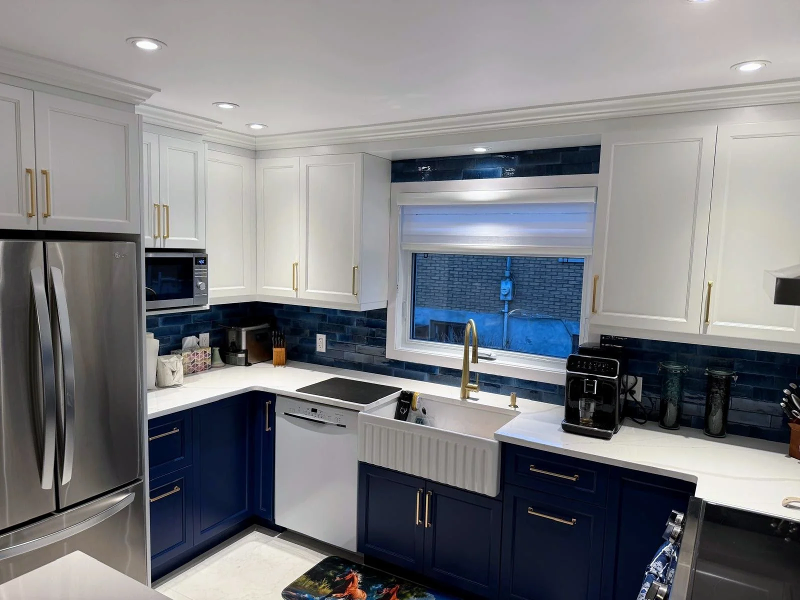 Modern kitchen with white upper cabinets, dark blue lower cabinets, white countertops, and stainless steel appliances. There is a window above the sink with a roll-up shade, and a black subway tile backsplash. The kitchen has recessed lighting.