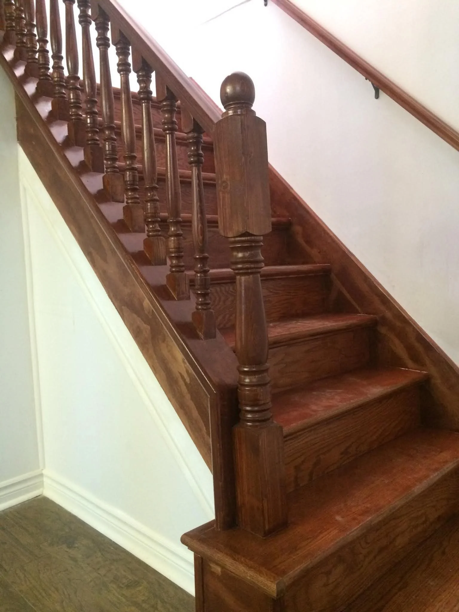 Wooden staircase with turned balusters and newel post, against white walls and a wooden floor.