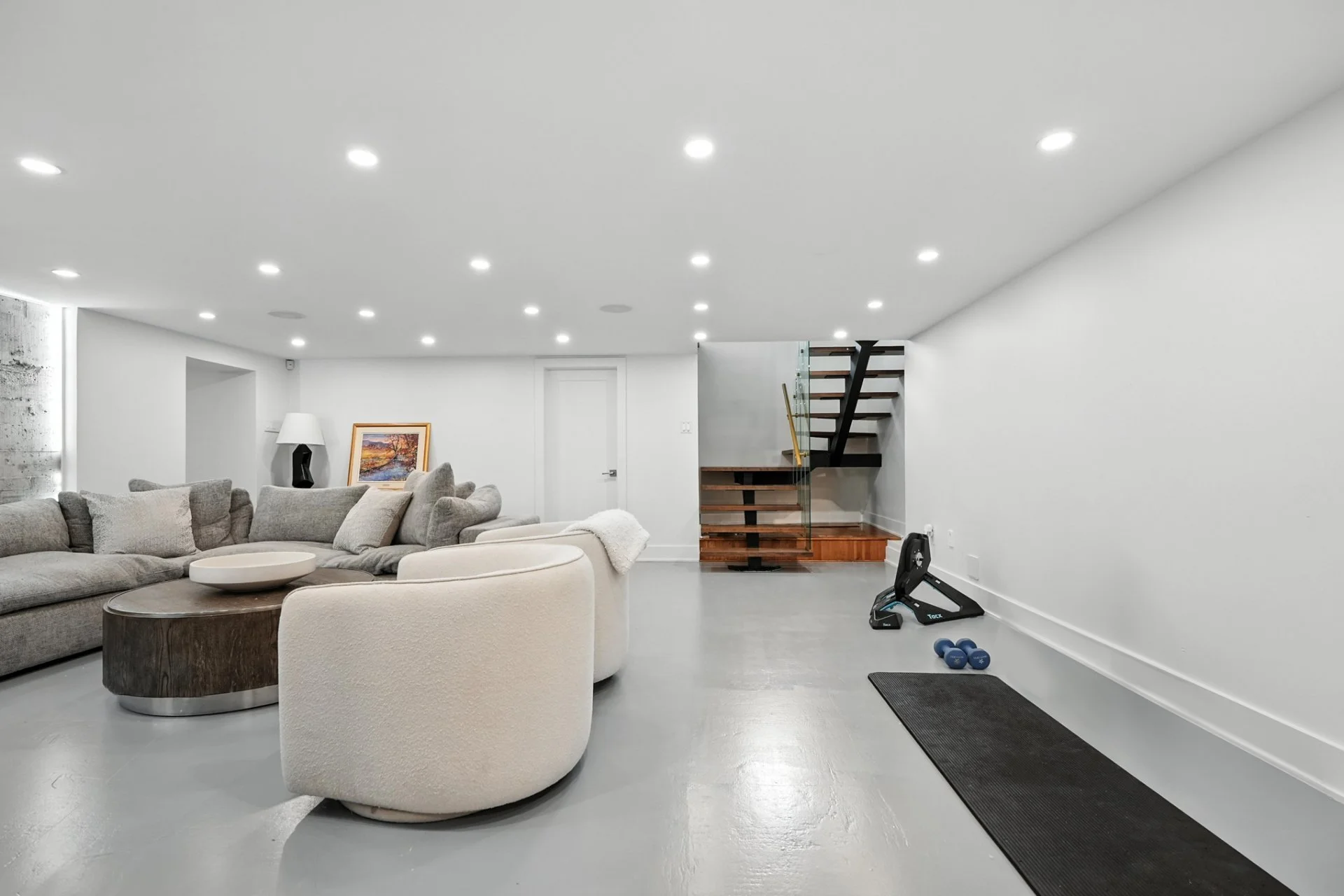 Modern living room with a white ceiling featuring numerous recessed lights, gray sectional sofa, white armchair, dark wooden coffee table, white lamp, framed landscape painting, treadmill, small dumbbells, black exercise mat, and a staircase with woo