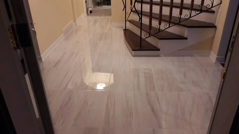 Indoor hallway with a shiny cream-colored tiled floor, a staircase with dark wooden steps and decorative iron railing, and walls painted light beige.