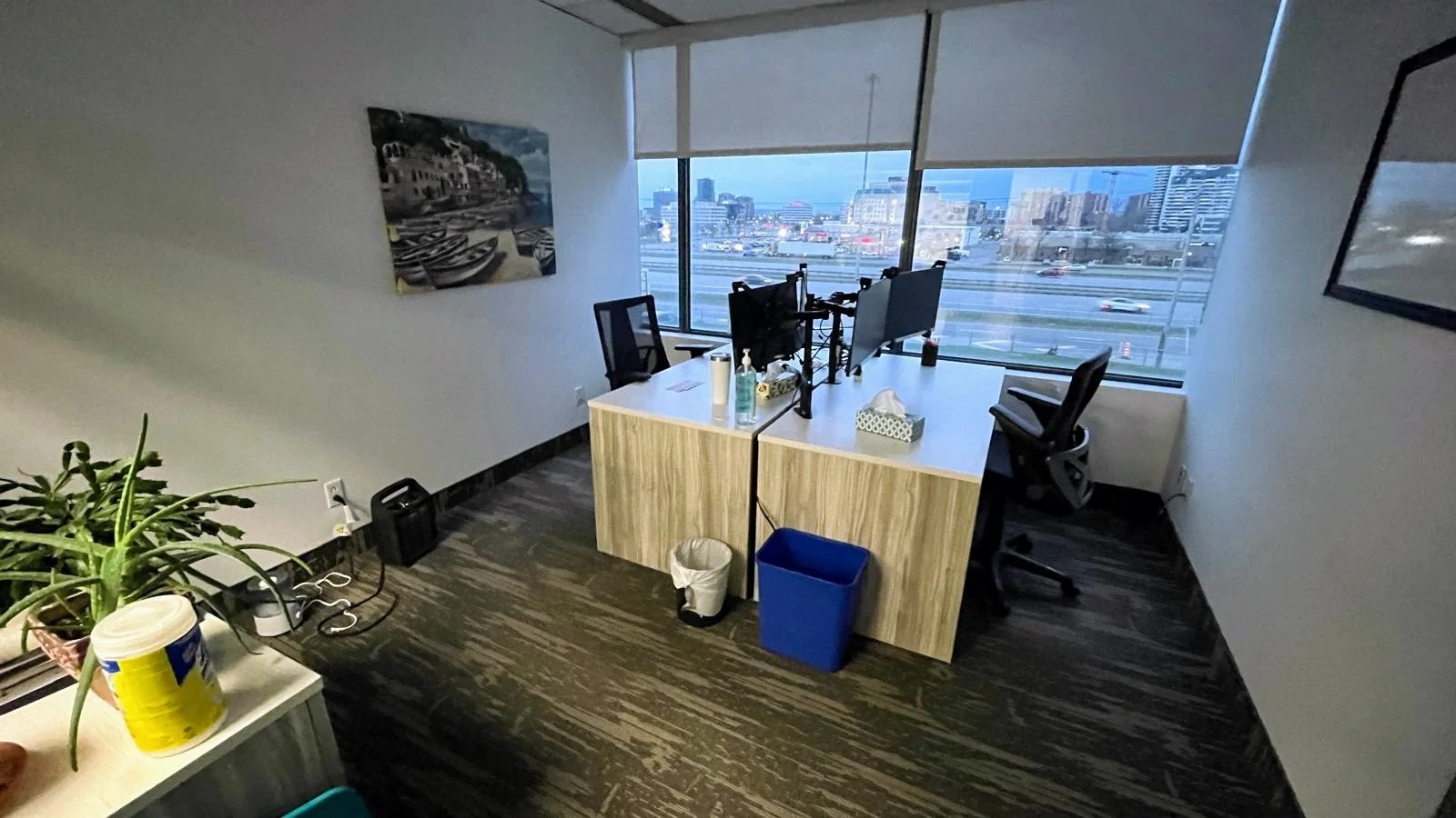 Office with two desks facing large window overlooking a city highway. One desk has dual monitors and office chairs, with supplies like tissues and sanitizer. The room has a painting on the wall and a plant on a side table.