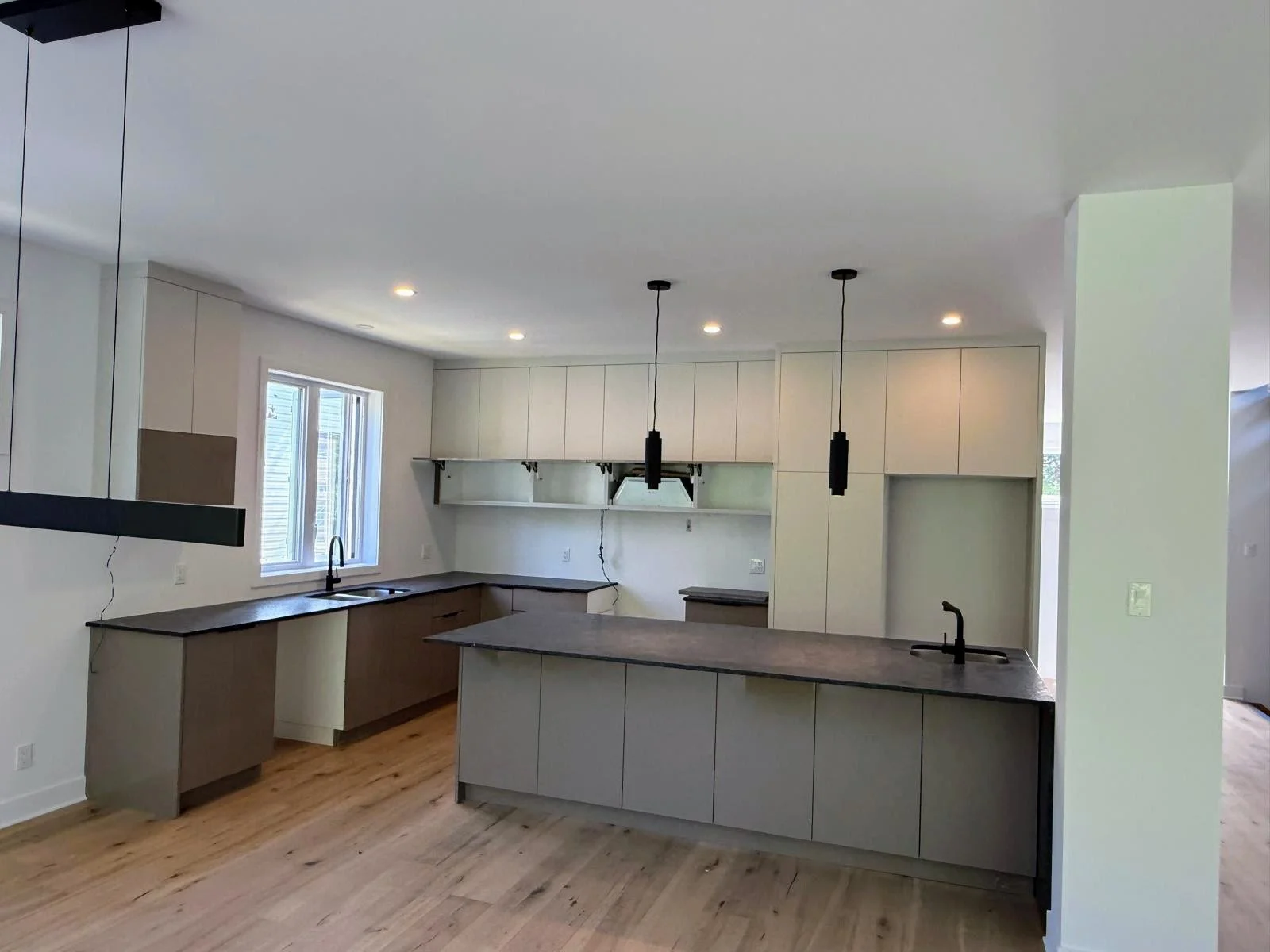 Modern kitchen with white cabinets, black countertops, wooden flooring, two black pendant lights hanging over the island, a window above the sink, and minimal decor.
