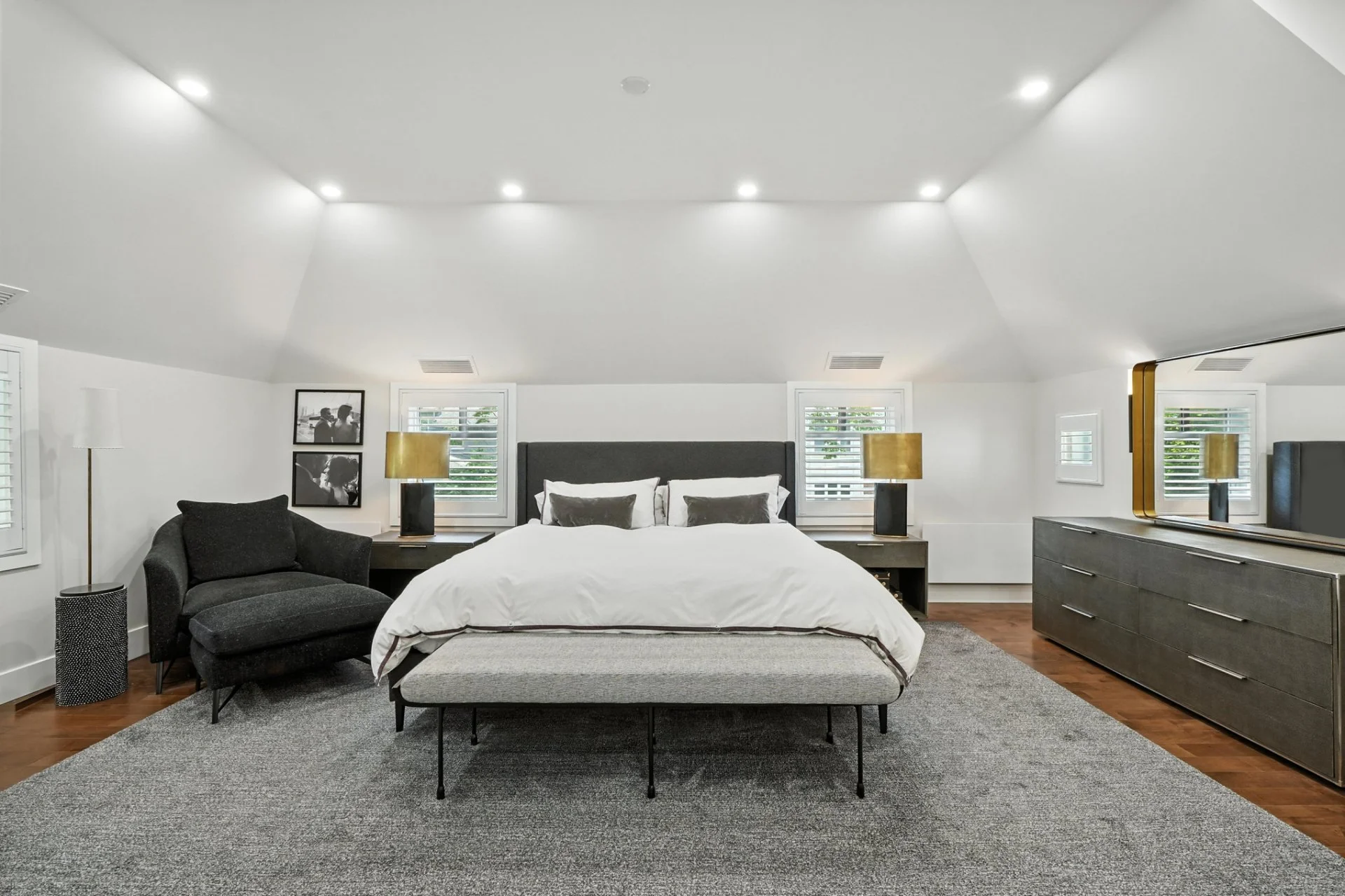 Modern bedroom with a large bed, gray headboard, white bedding, black and gray pillows, two nightstands with gold lamps, a black armchair, framed black-and-white photographs, a gray dresser with a mirror, and wooden floors with a gray area rug.
