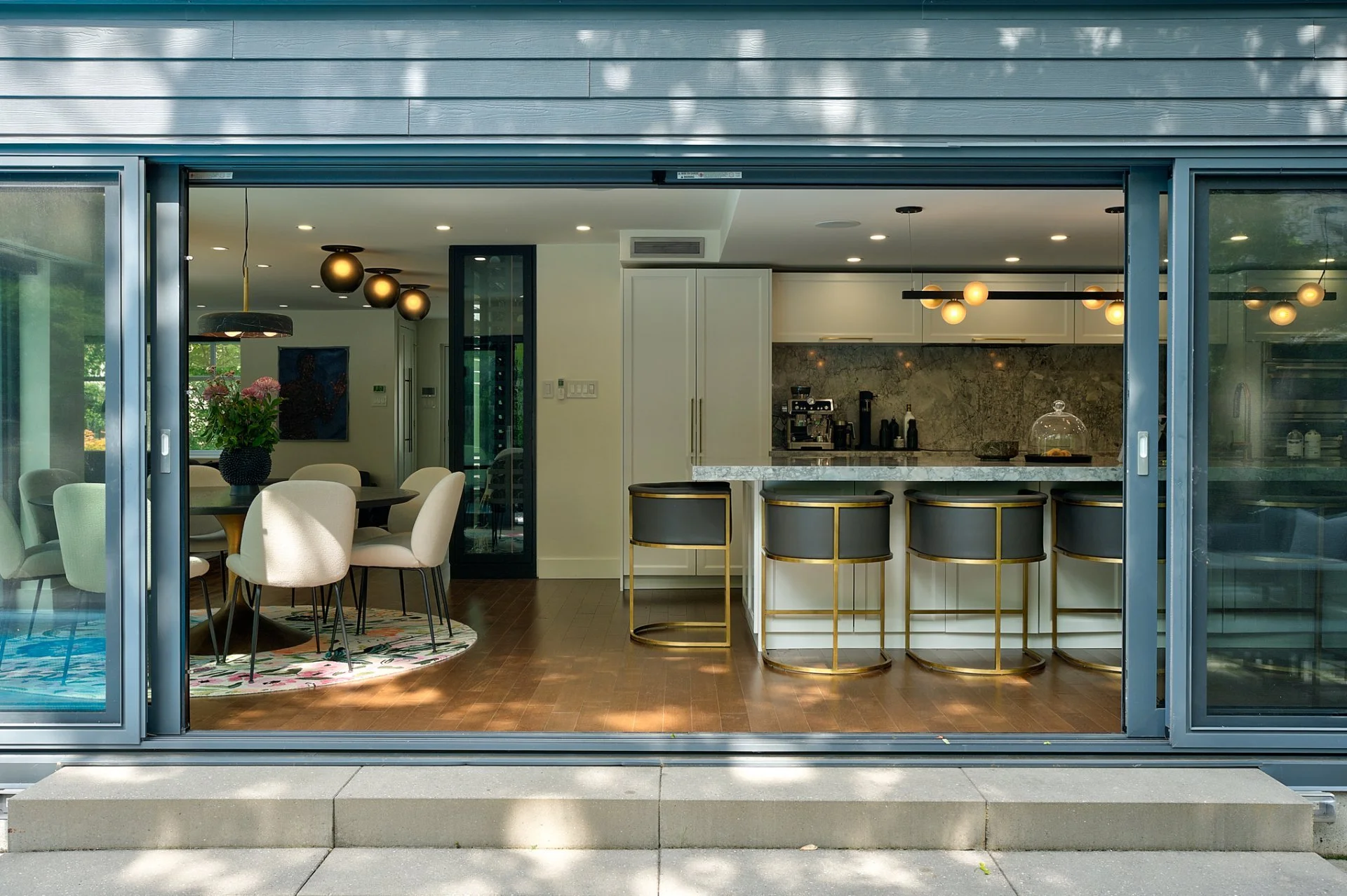 Interior view of a modern kitchen and dining area seen through sliding glass doors, with a kitchen island, bar stools, and a dining table with chairs.