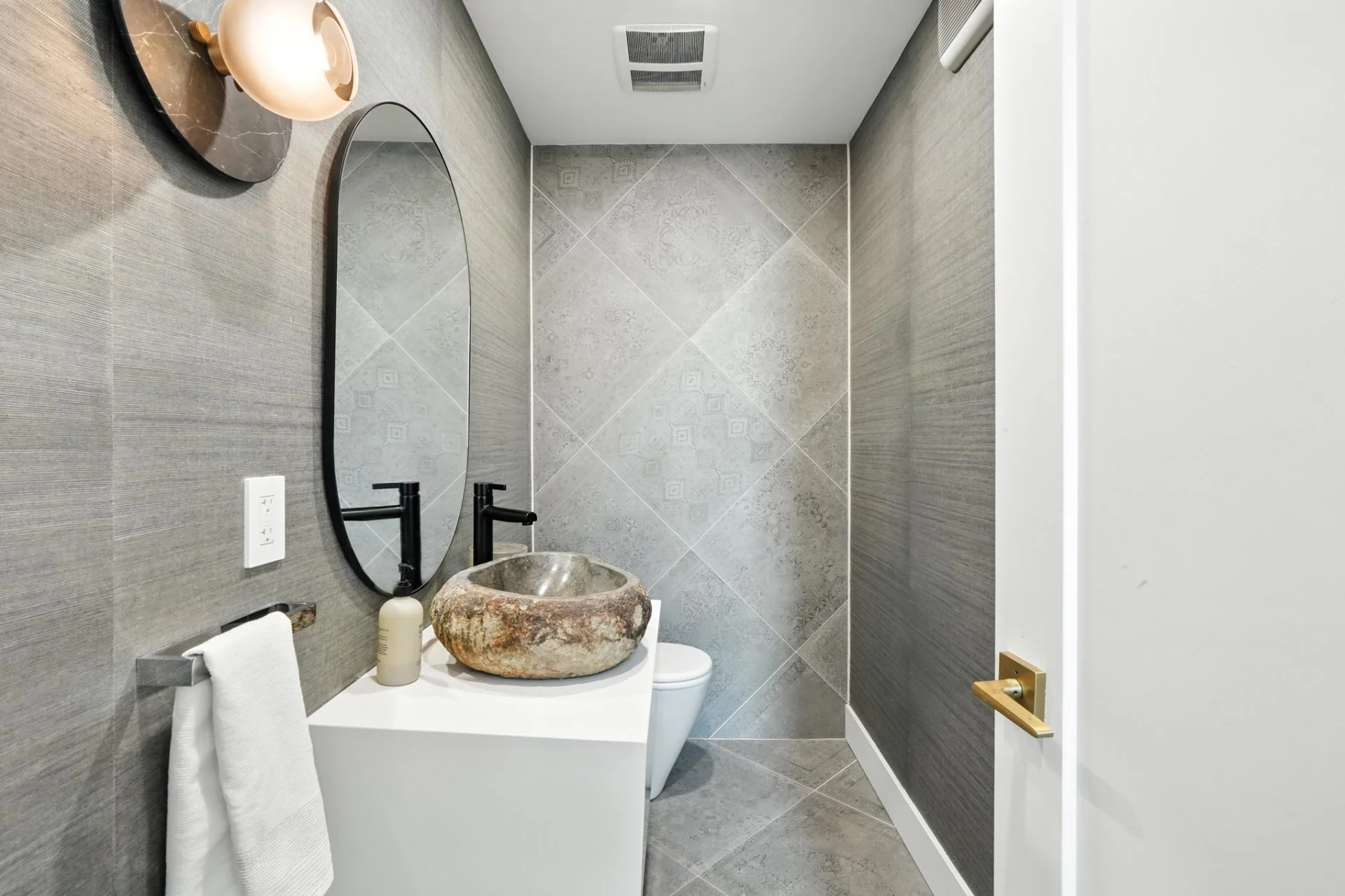 Modern bathroom with gray textured wall and large diagonal gray tiles on the back wall, a white vanity with a stone vessel sink, black faucet, oval mirror, hand towel on a rail, and a decorative wall-mounted light fixture.