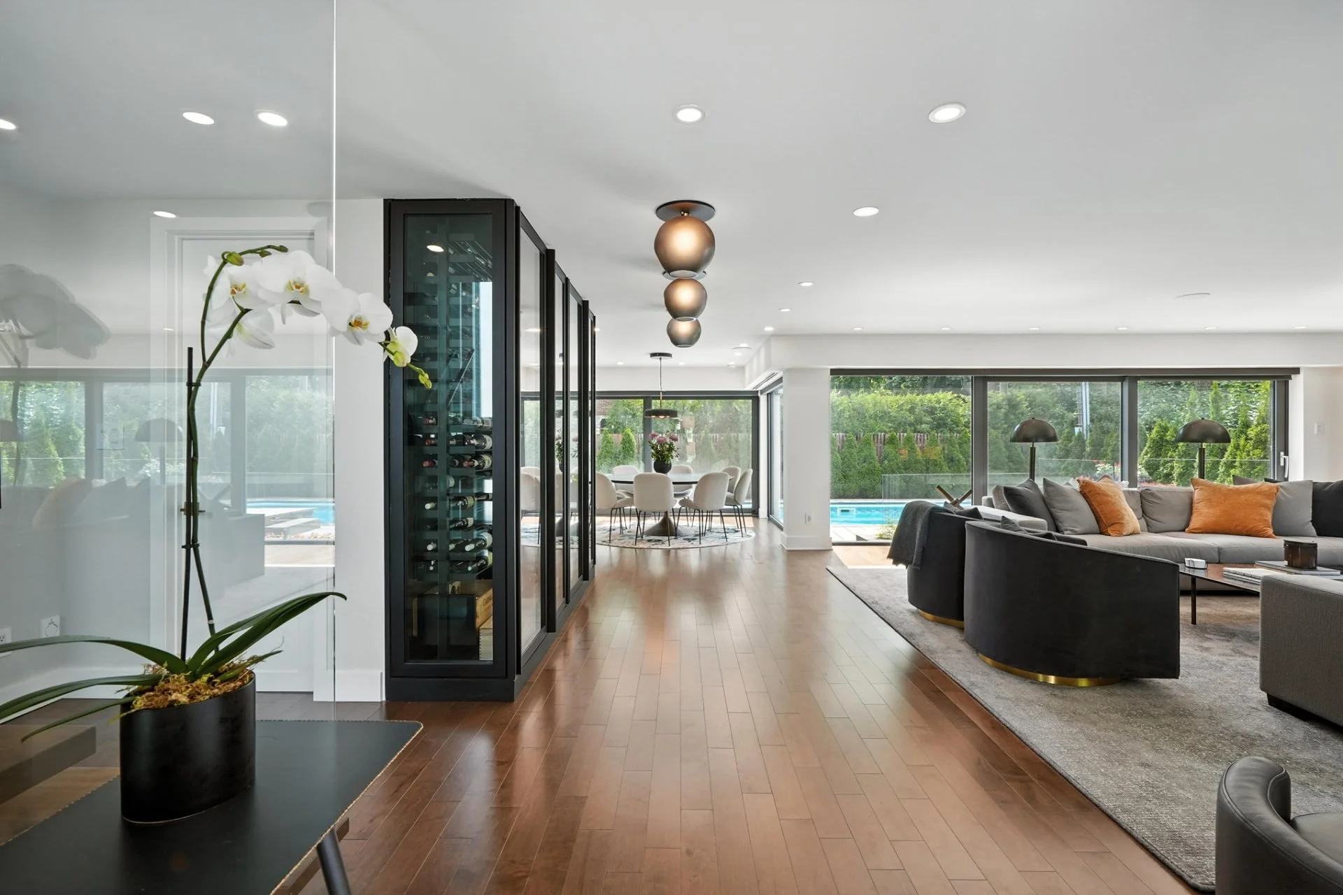 Modern living room with hardwood floors, large windows, black sectional sofa with orange pillows, large gray area rug, wine fridge, and view of backyard pool.