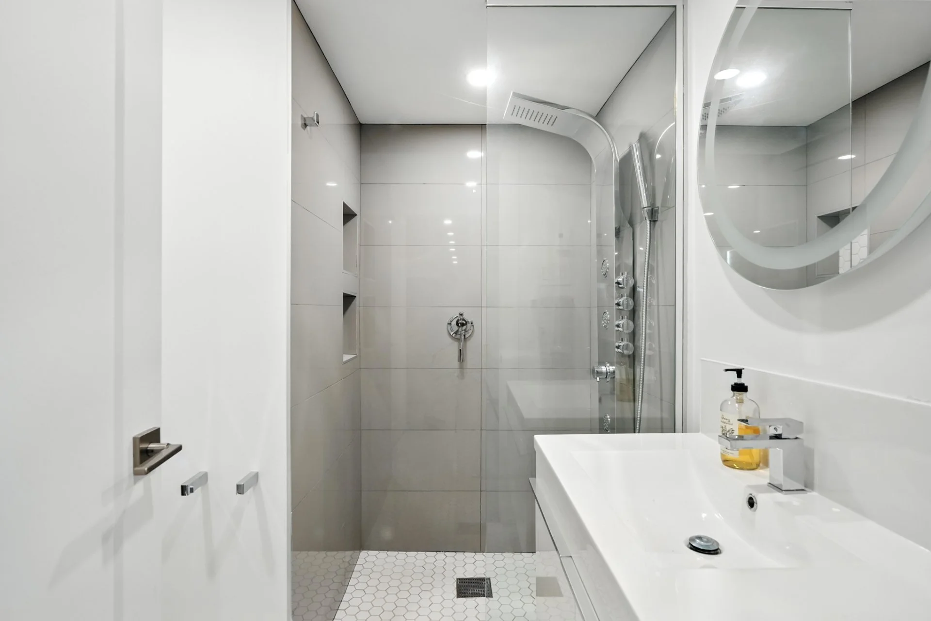 Modern bathroom with white walls, a large mirror, a white sink, a bottle of soap, and a shower area with gray tiles and a glass door.
