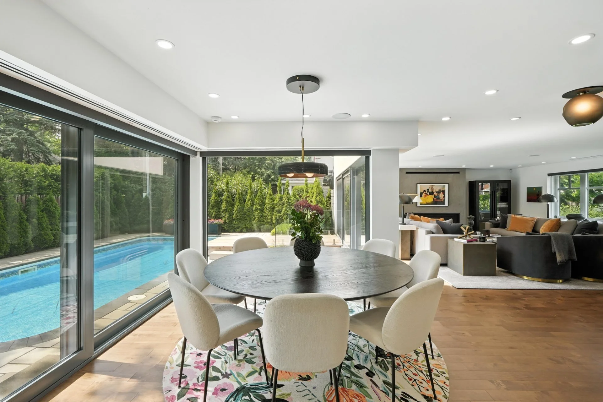 Interior view of a modern open-plan home with a dining area, living room, and backyard swimming pool visible through large glass sliding doors, with greenery outside.