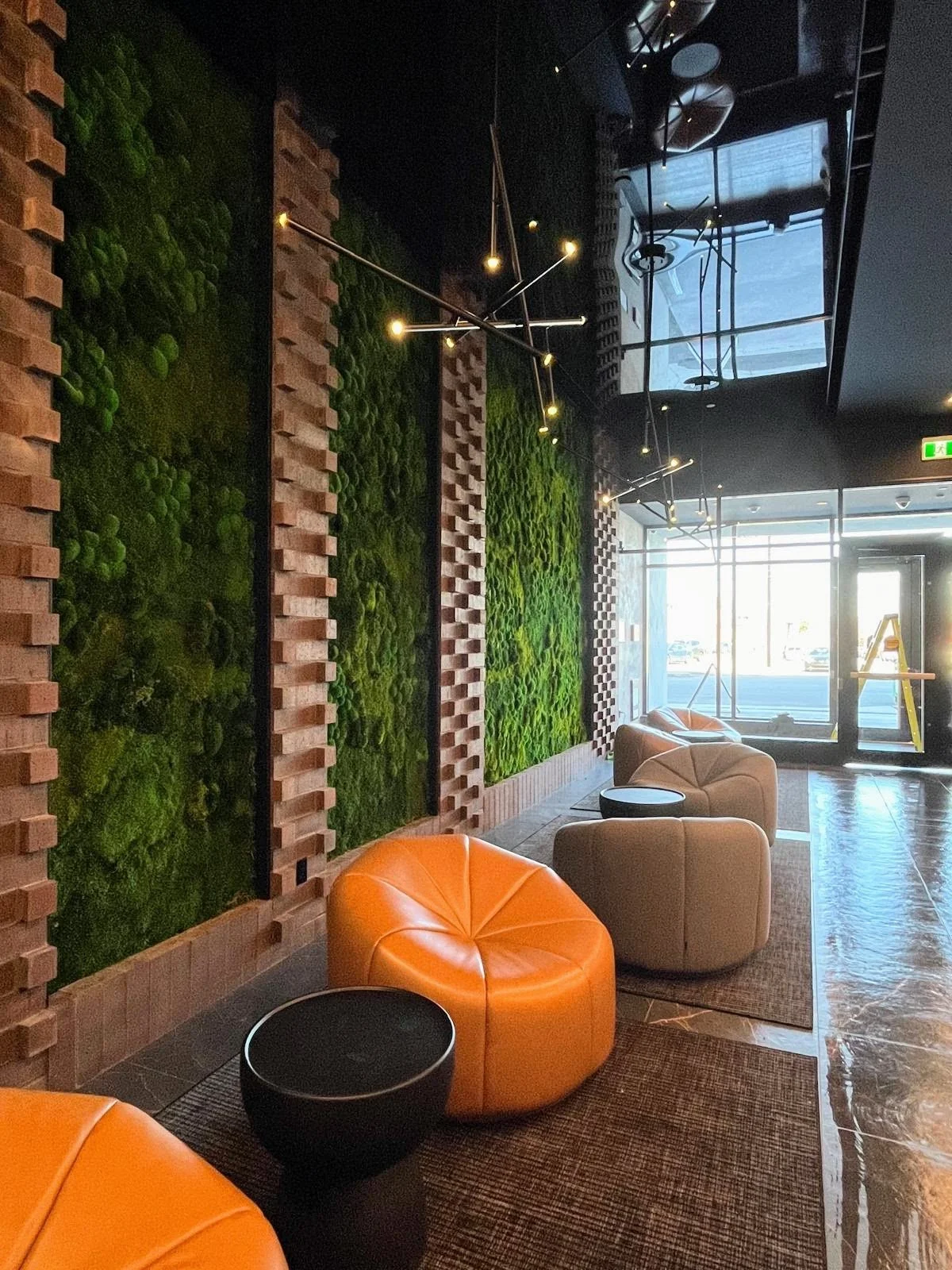 Modern lobby with brick and moss wall, orange and beige chairs, black side table, and hanging light fixtures, with large glass windows and door at the entrance.