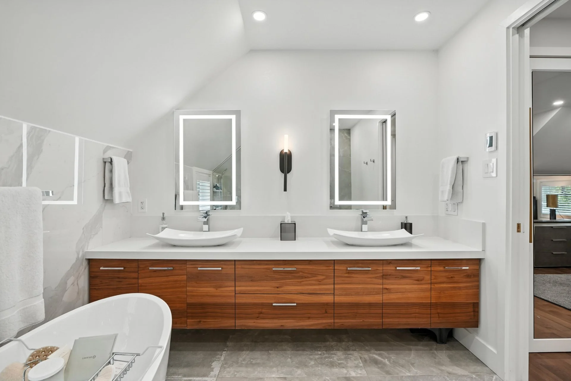 Modern bathroom with double vanity, two rectangular mirrors with LED borders, and vessel sinks. The vanity has wood cabinets and a white countertop. A wall sconce and towels are also visible.