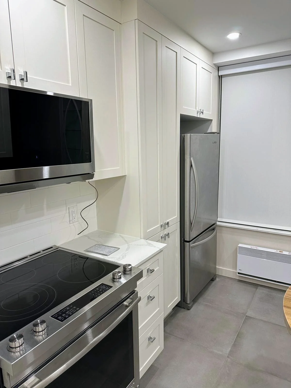 Modern kitchen with white cabinets, stainless steel refrigerator, black electric stove, microwave, white countertop, and window with roller shade.