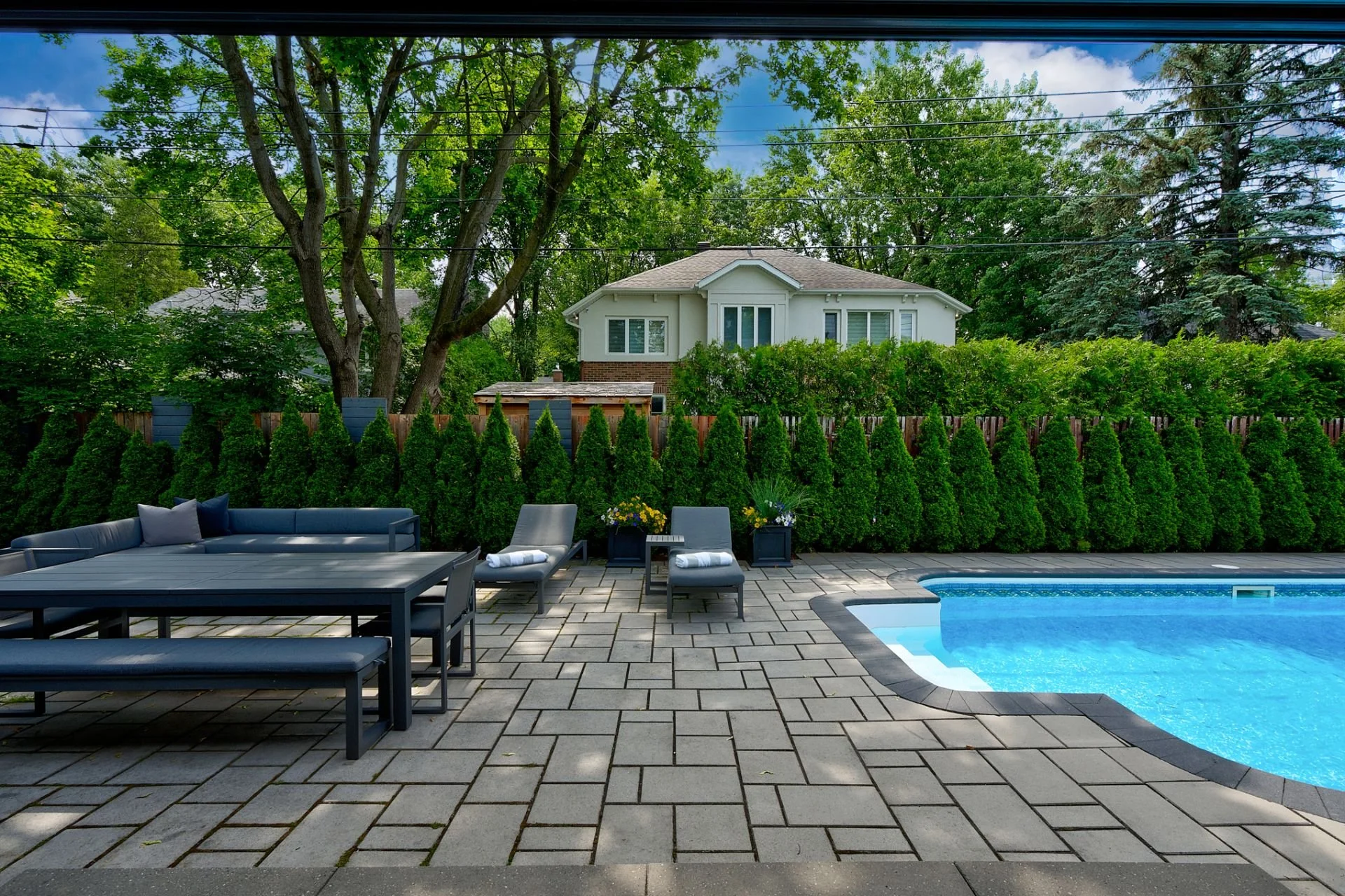 Backyard with swimming pool, gray outdoor furniture, potted plants, lush green bushes, tall trees, and a white house in the background.