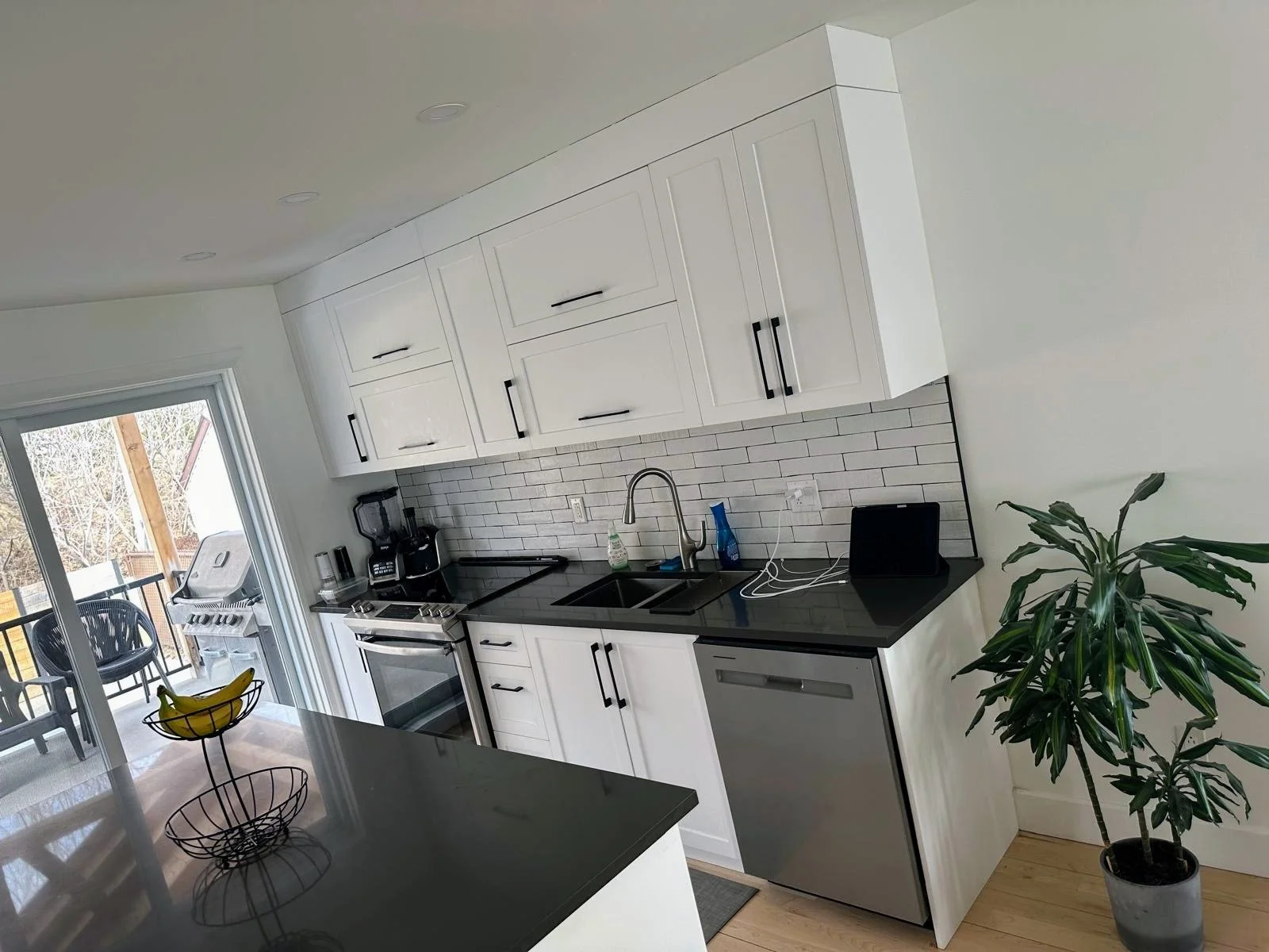 Modern kitchen with white cabinets, black countertop, stainless steel dishwasher, and a sliding glass door leading to an outdoor balcony with a grill and chairs. There is a potted plant on the right and various small appliances on the counter.