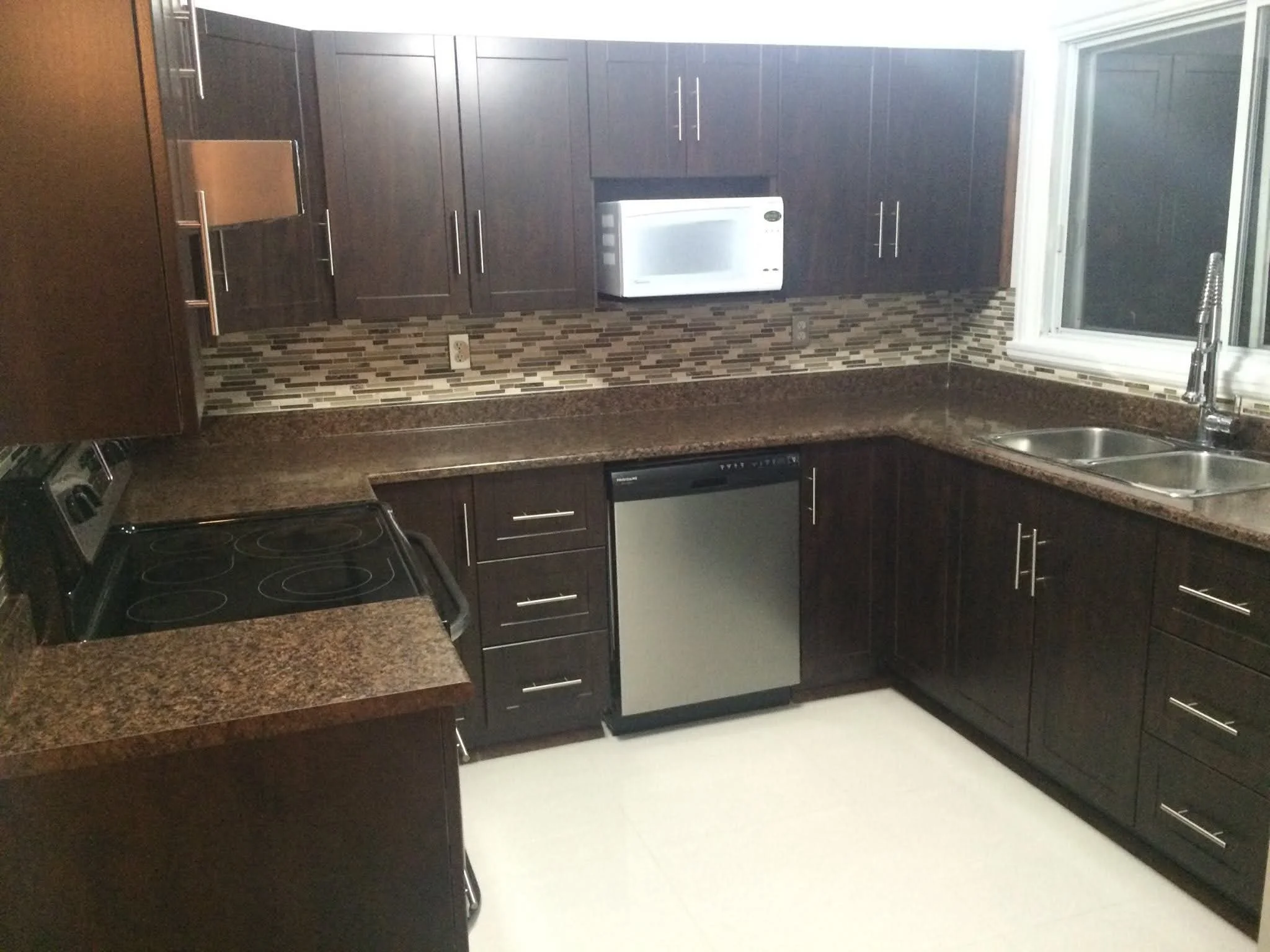 Kitchen with dark wood cabinets, granite countertops, mosaic tile backsplash, stainless steel microwave, electric stove, dishwasher, and double sink near a window.