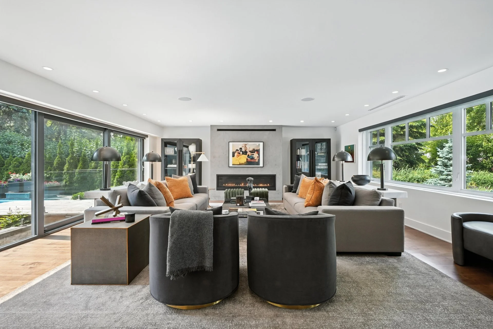 Modern living room with large windows, two sofas with pillows, a fireplace, artwork above fireplace, bookshelves, black lamps, and outdoor greenery visible through windows.