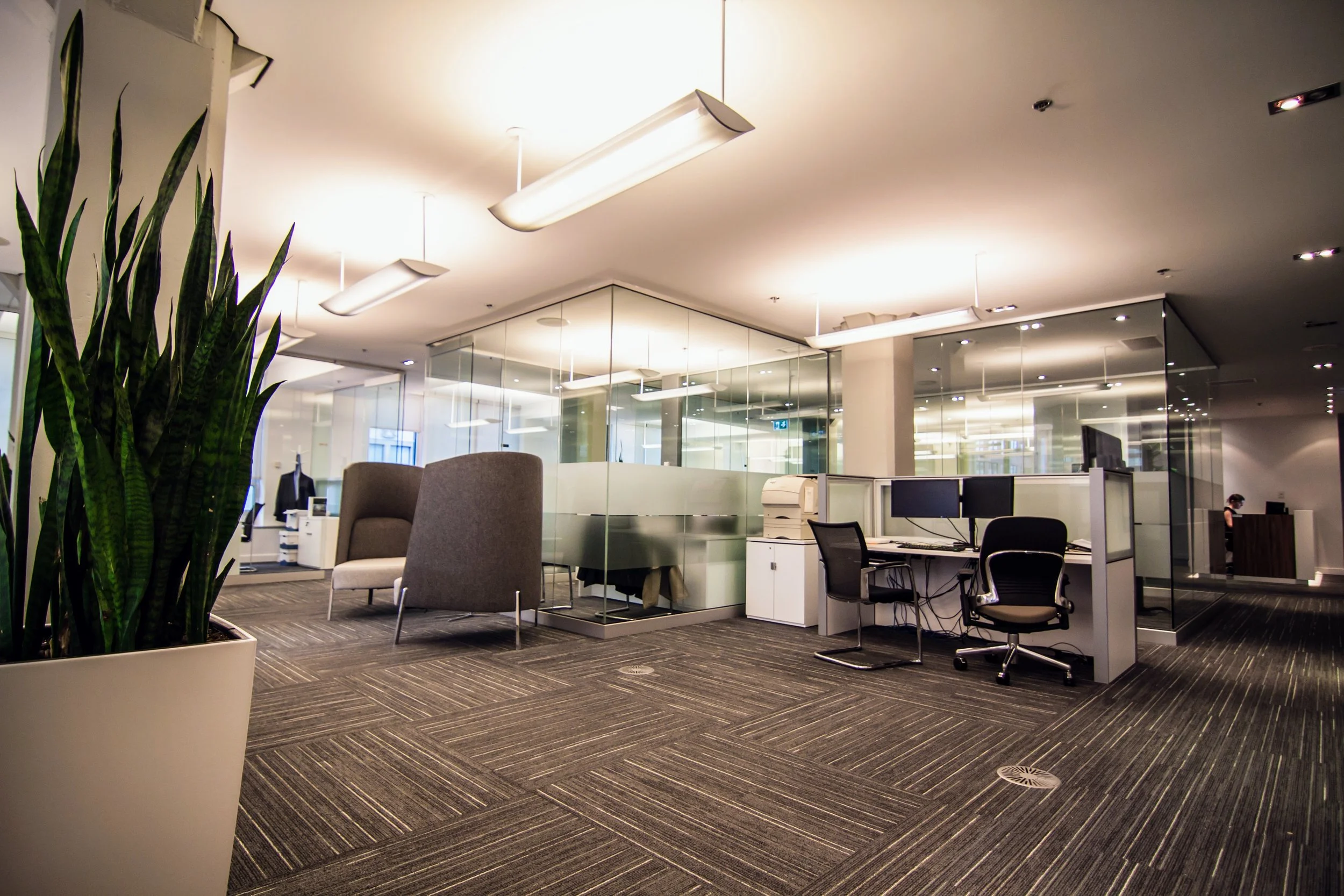 Modern office space with glass-walled meeting rooms, desks with computer monitors, office chairs, and potted plants, illuminated by ceiling lights.
