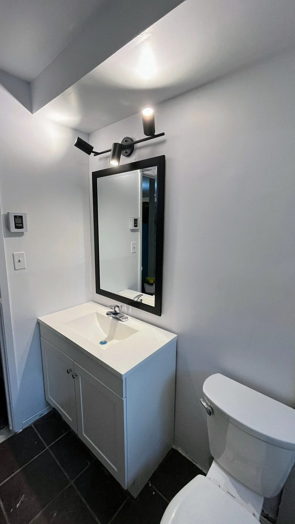 A small bathroom with a white sink vanity, black framed mirror, wall-mounted light fixture, and a white toilet, with black tile flooring.