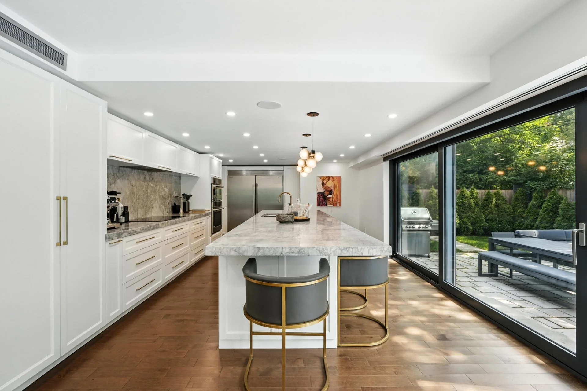 Modern kitchen with white cabinets, marble countertops, an island with two bar stools, and large sliding glass doors opening to a backyard with outdoor seating and greenery.